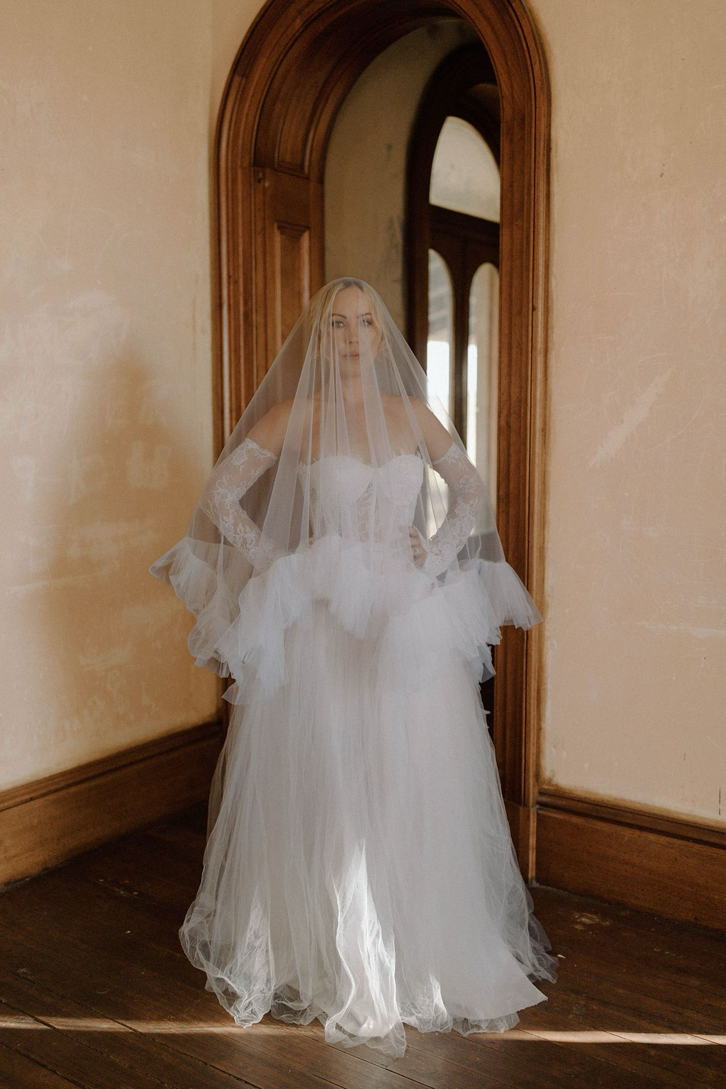 A woman in a wedding dress and veil standing in front of a mirror in a room with wooden trim and beige walls.