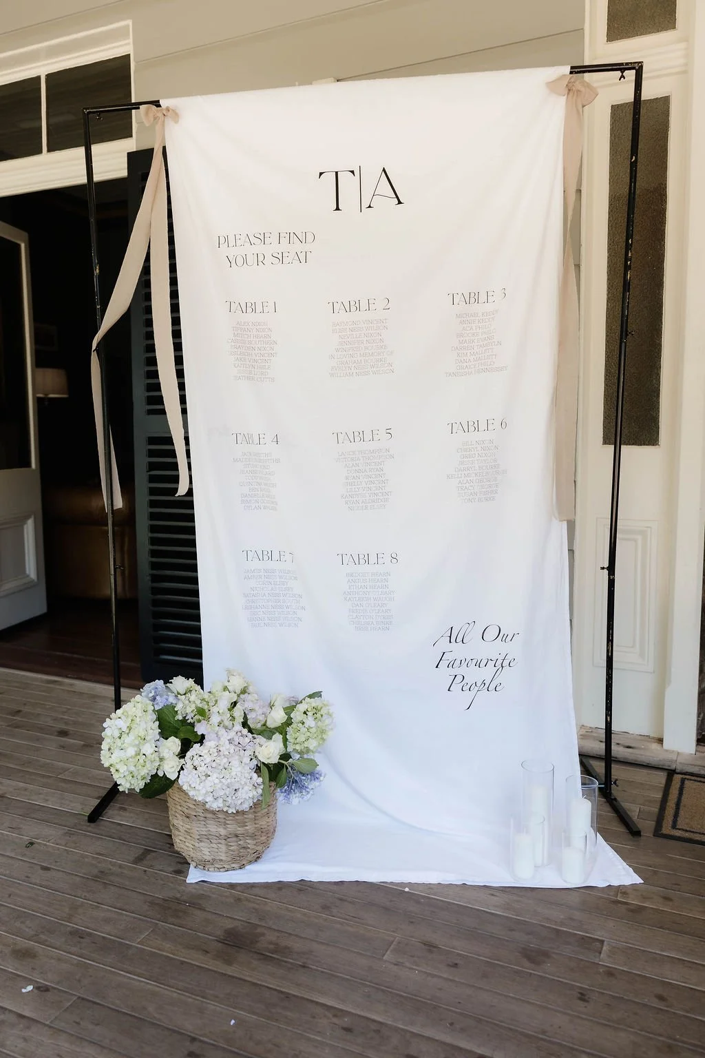 Wedding seating chart with table numbers and guest names on a white fabric backdrop, decorated with white and lavender flowers in a basket, and white candles on the floor. Toowoomba wedding Gabbinbar homestead 