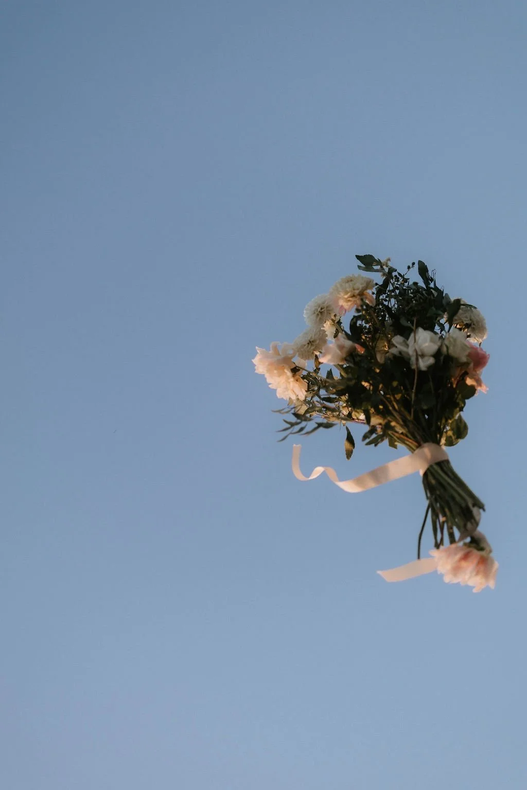 A bouquet of white and light pink flowers with green leaves tied with a white ribbon, held up against a clear blue sky.