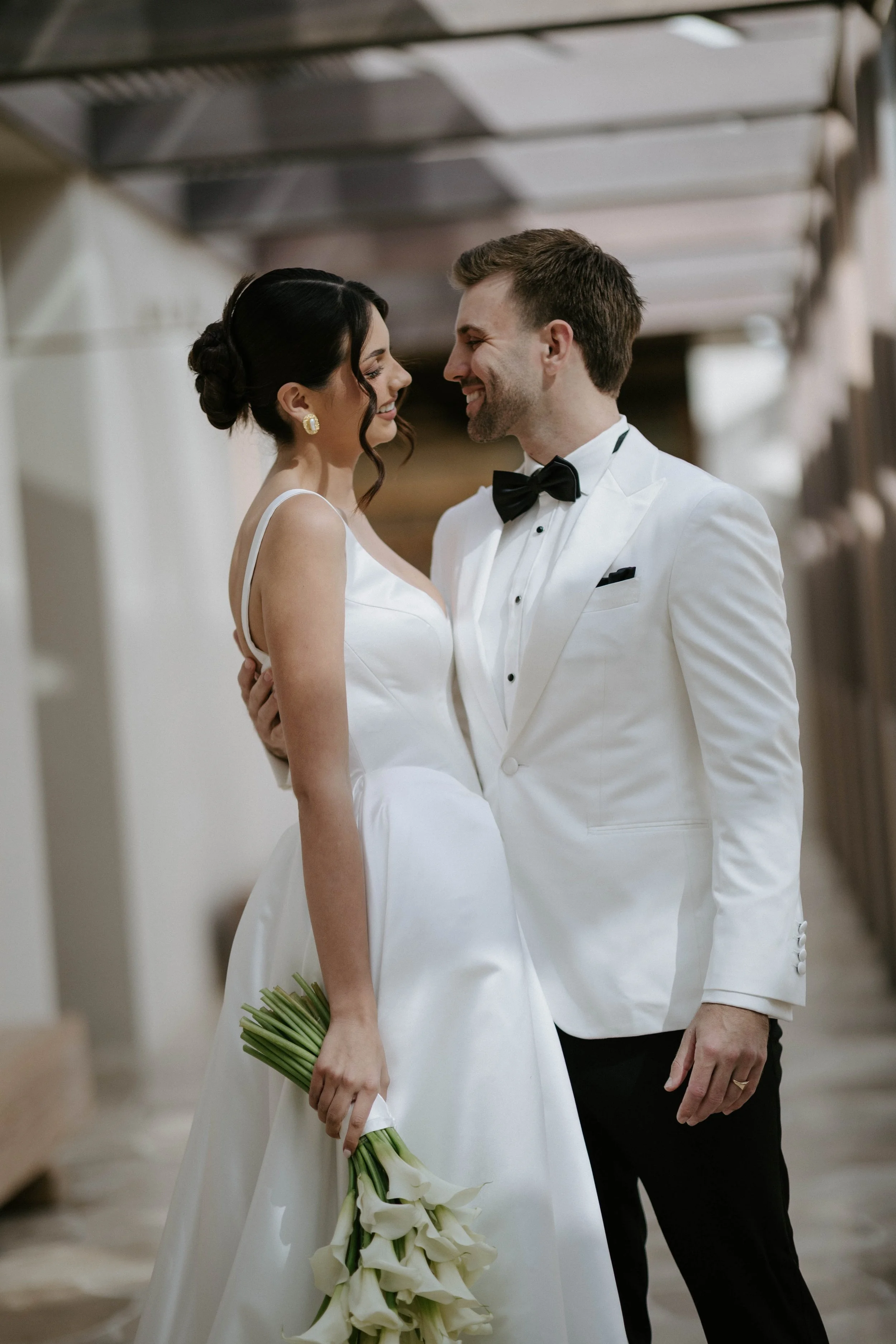 A bride and groom smiling closely in wedding attire, with the bride holding a bouquet of white calla lilies, indoors with industrial ceiling elements in the background.