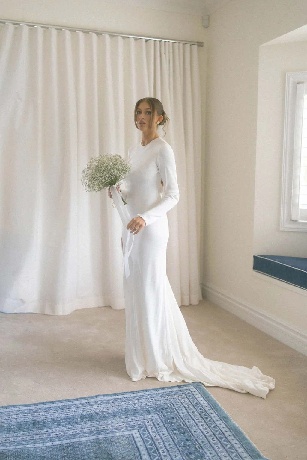 A woman in a white wedding gown holding a bouquet of baby's breath flowers in a room with white curtains and a window.