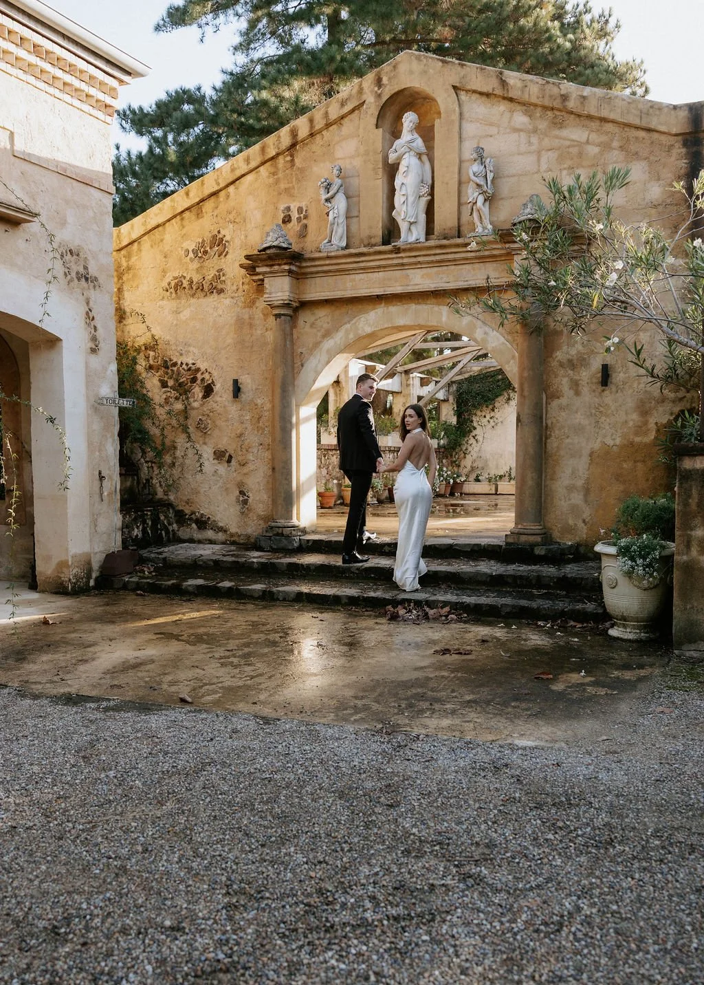A couple dressed in wedding attire standing on steps underneath an arched structure with statues on top, in a courtyard with a vintage, rustic appearance. NSW wedding photographer Byron Bay 