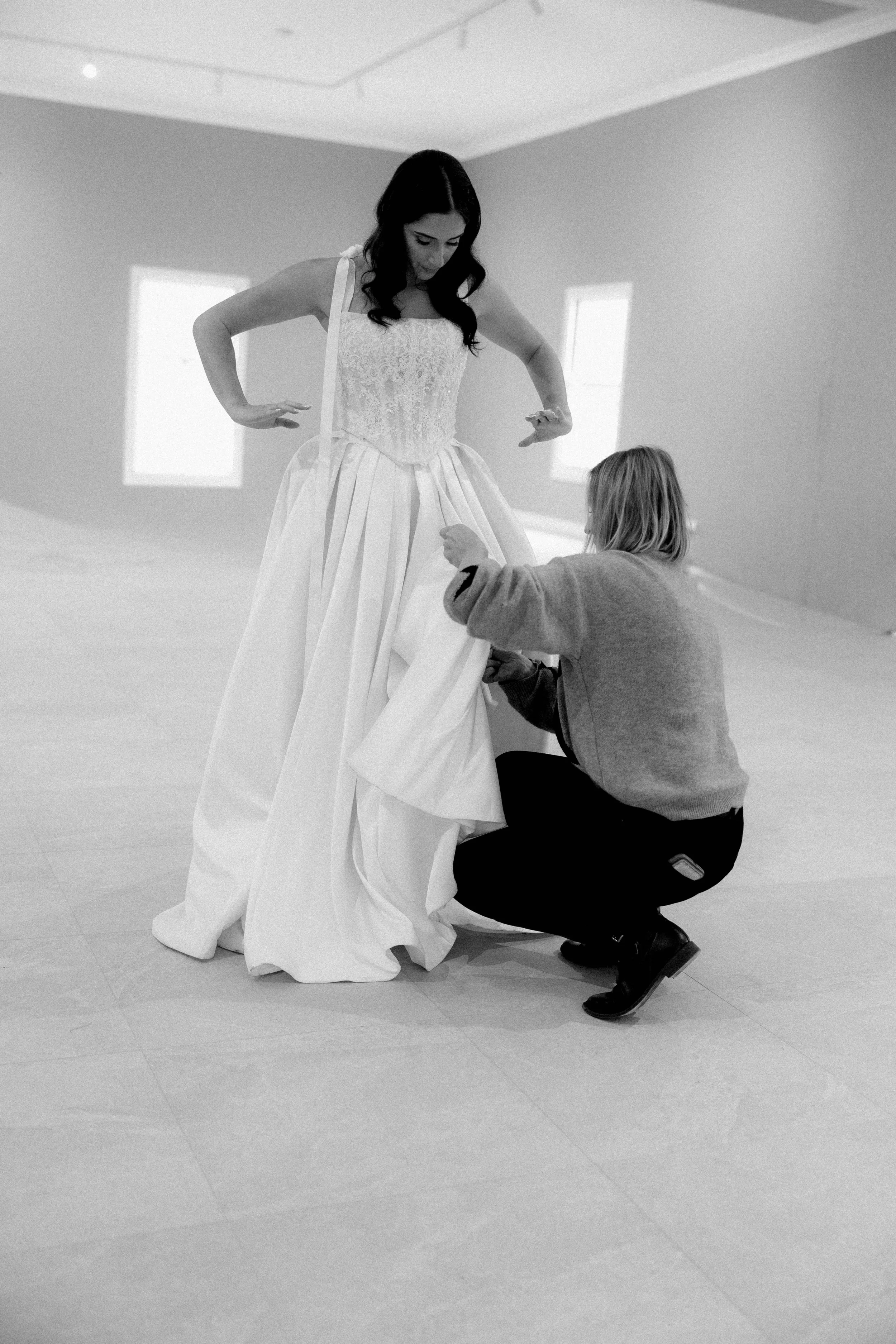 Woman in a wedding dress being assisted by a person crouching down, possibly a designer or seamstress, in a minimalist room with two small windows.
