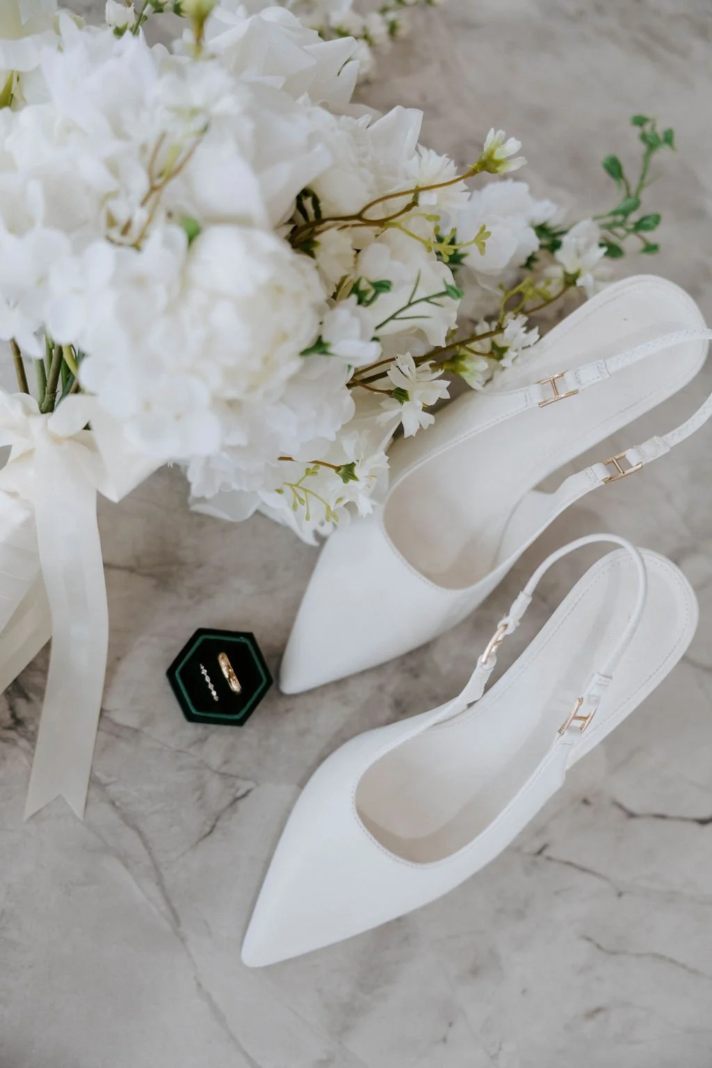 White high-heeled shoes with ankle straps, a bouquet of white flowers, and a black ring box with a gold ring inside on a marble floor.