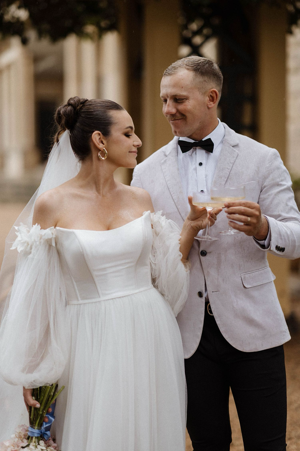 A bride and groom sharing a glass of champagne at their wedding celebration. The bride is wearing a white wedding gown, and the groom is dressed in a light-colored suit with a bow tie.