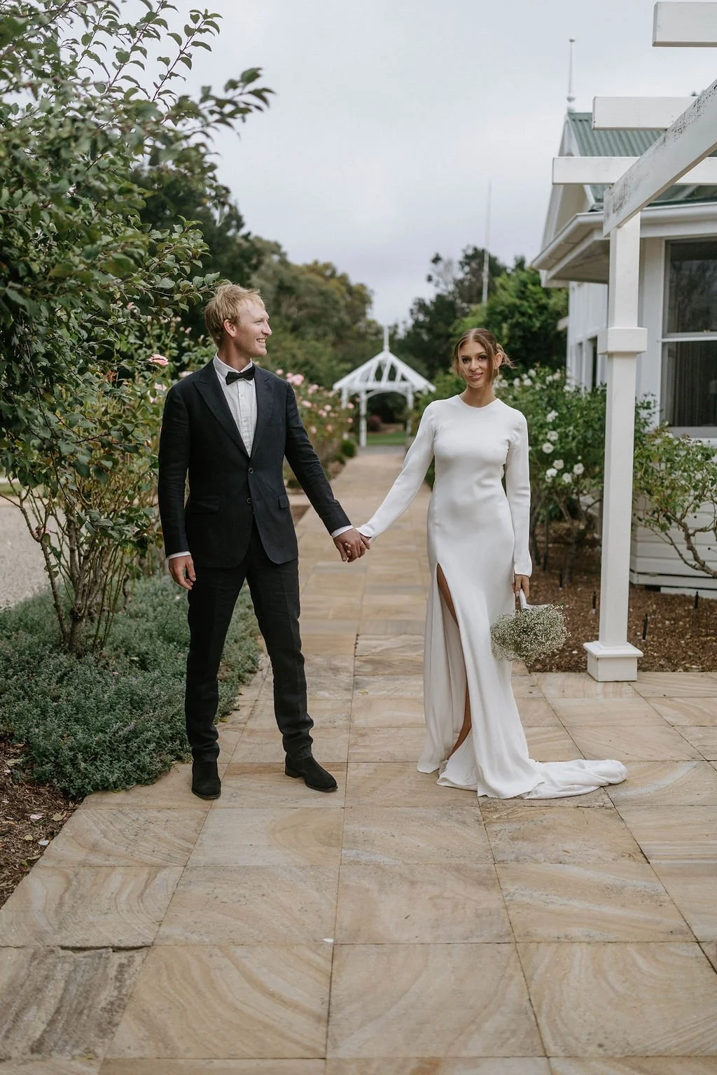 A newlywed couple holding hands outdoors on a stone pathway, with the groom in a black tuxedo and the bride in a long white wedding dress holding a bouquet of baby's breath flowers. They are smiling at each other near a white house with garden bushes