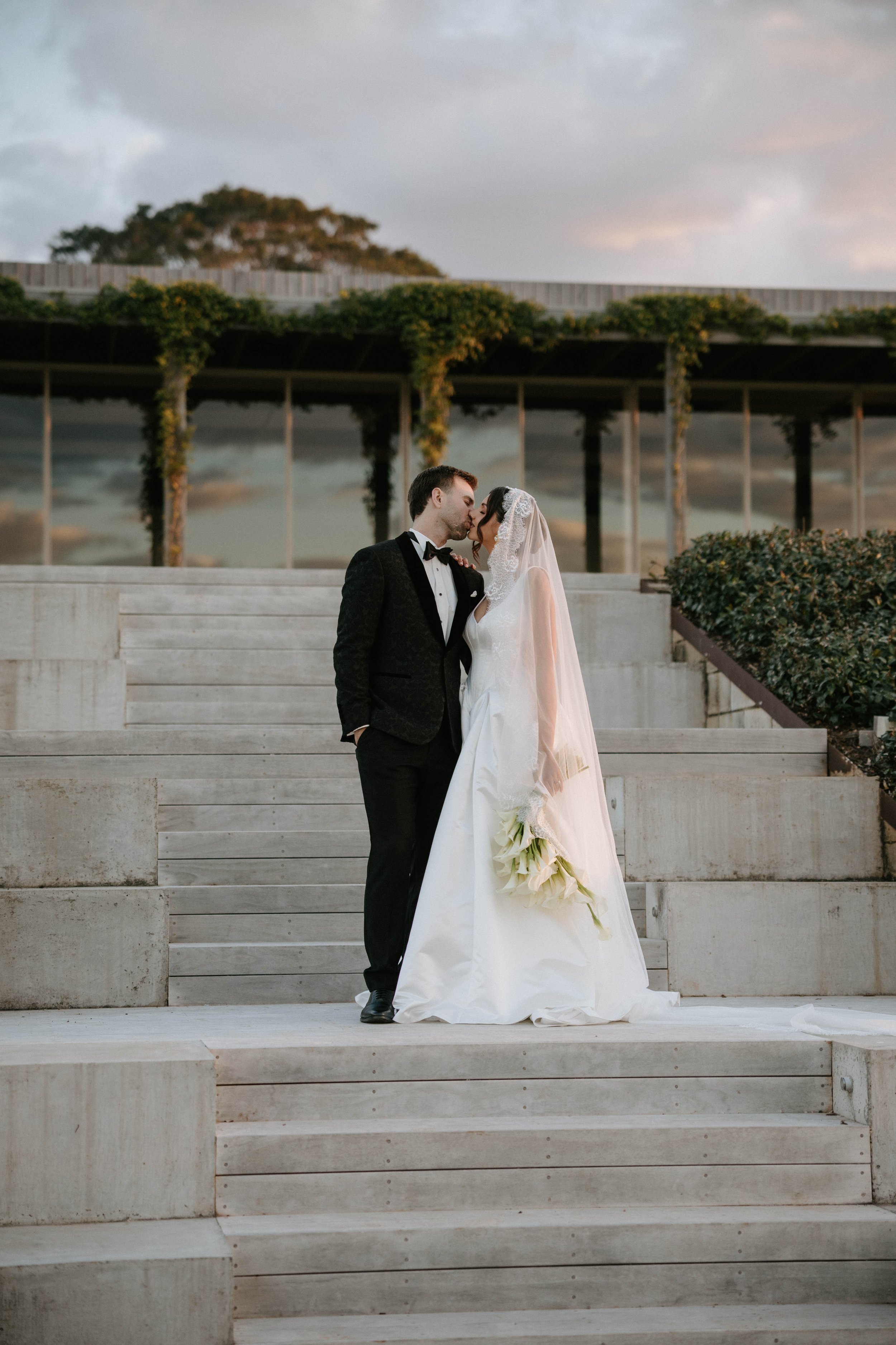 A bride and groom kissing on the steps of an outdoor location, with the bride holding a bouquet, greenery climbing a structure behind them, and a partly cloudy sky in the background during sunset.