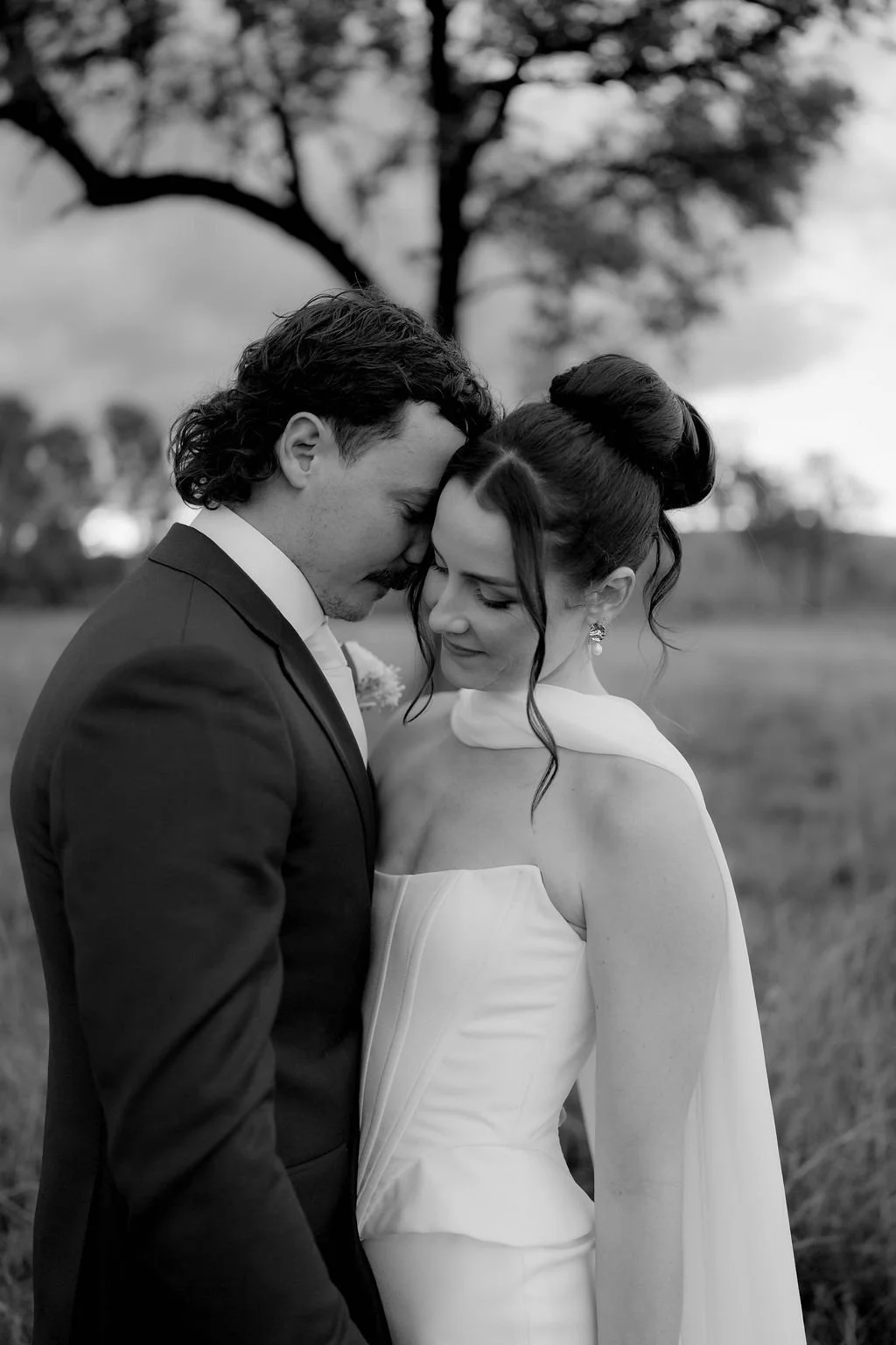 A black and white photo of a bride and groom standing close, with their foreheads touching and eyes closed, outdoors with a tree and cloudy sky in the background.
