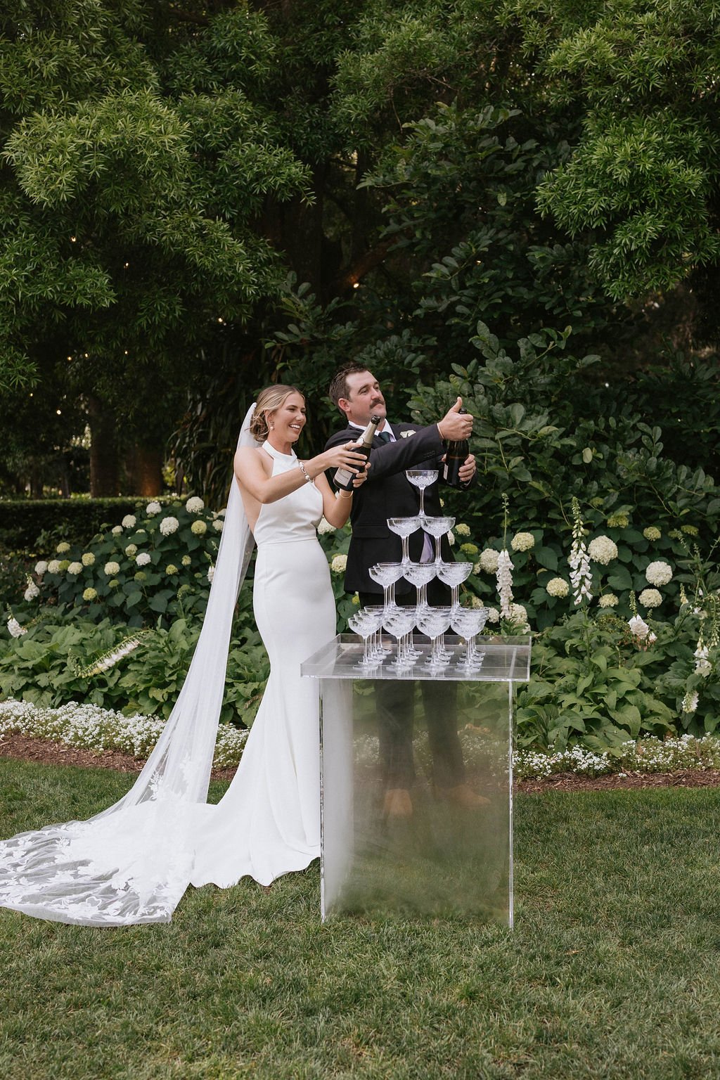 A bride and groom in wedding attire celebrating outdoors by opening a champagne bottle into a tower of empty champagne glasses, with lush green trees and flowering bushes in the background.