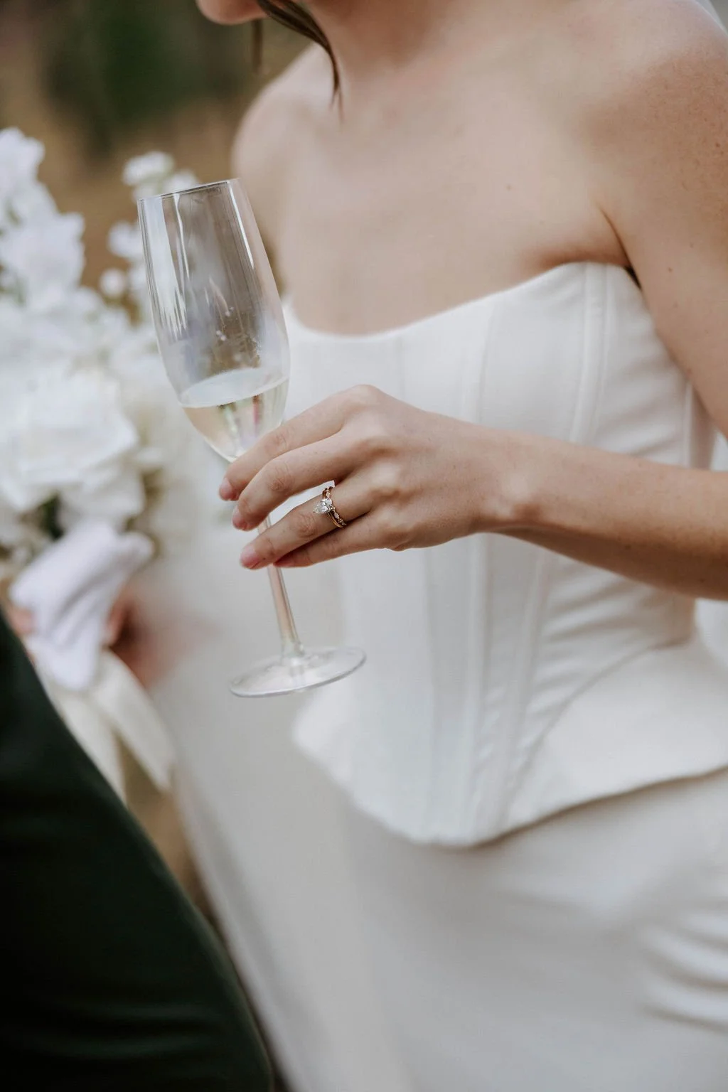 A bride holding a glass of champagne during her wedding ceremony, wearing a white strapless wedding dress and a ring on her finger.