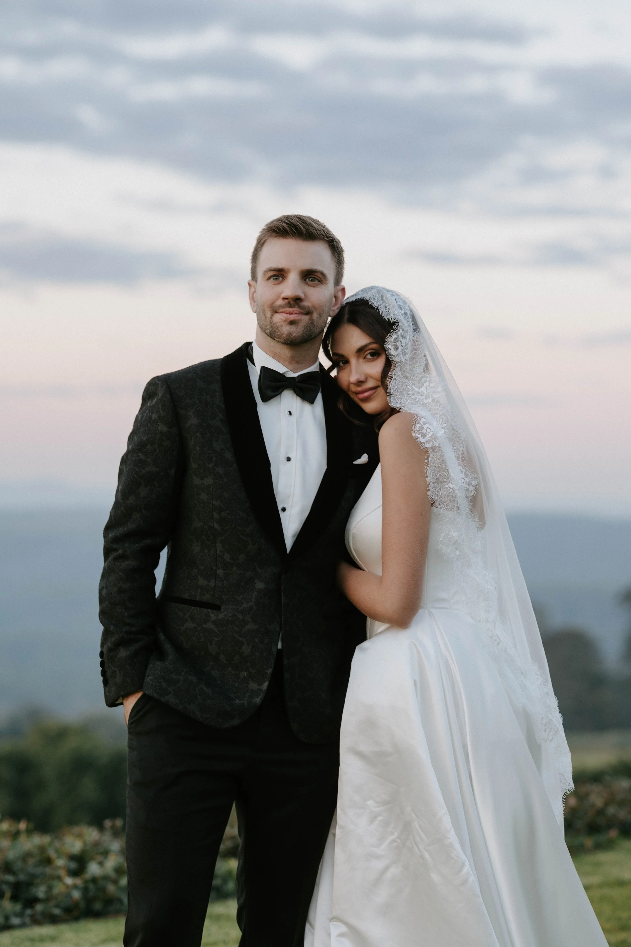 A bride and groom stand together outdoors against a cloudy sky. The groom is wearing a black tuxedo with a bow tie, and the bride is wearing a white wedding dress with lace details and a veil. They are close, with the bride leaning her head on the gr
