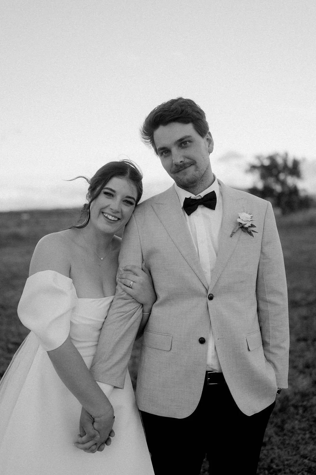 Black and white photo of a smiling woman in a white off-shoulder wedding dress holding hands with a man in a light suit and bow tie, standing outdoors with a blurred landscape and tree in the background. wedding photographer Brisbane Toowoomba