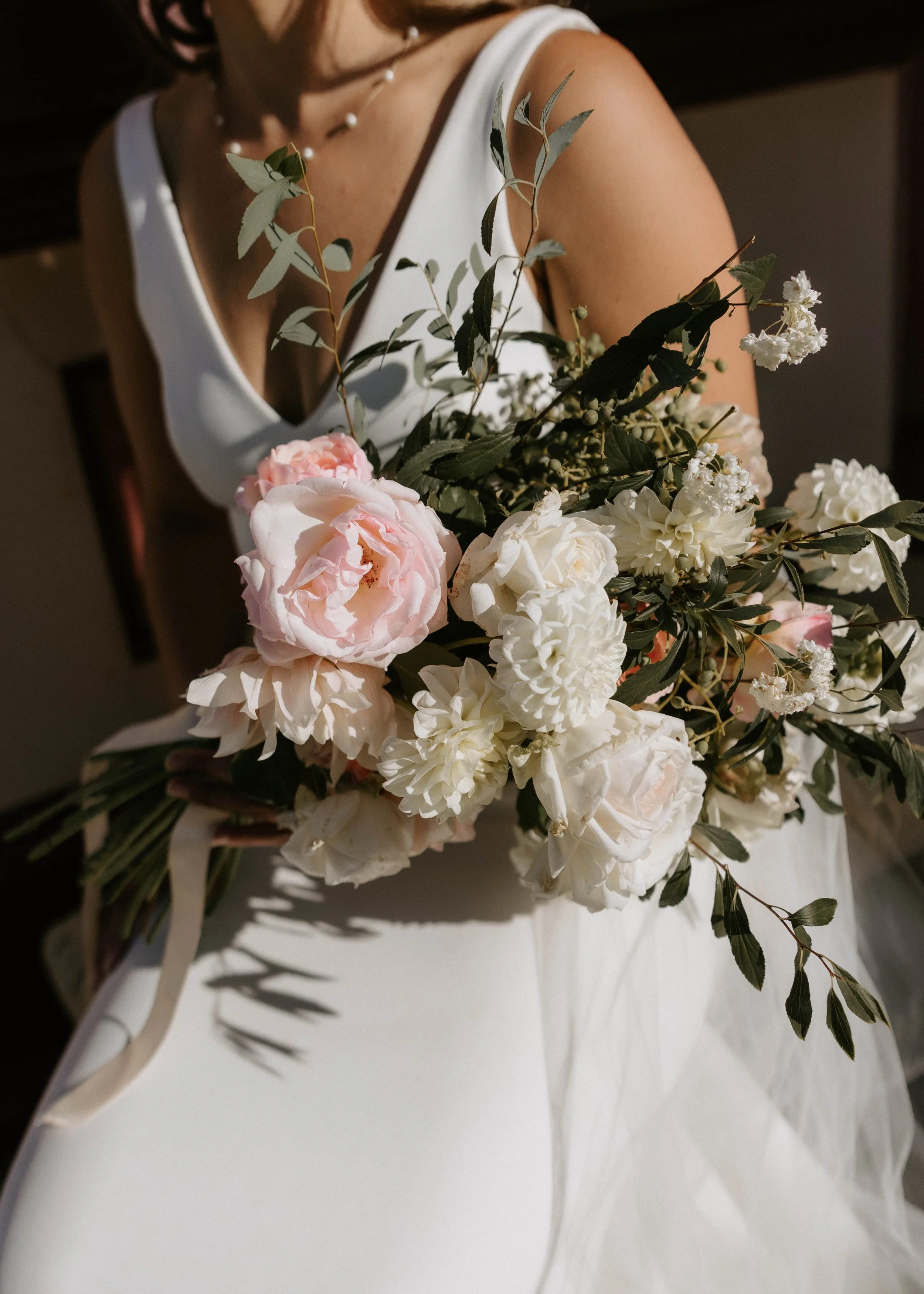 Close-up of a woman in a white dress holding a bouquet of white and pale pink flowers and greenery.