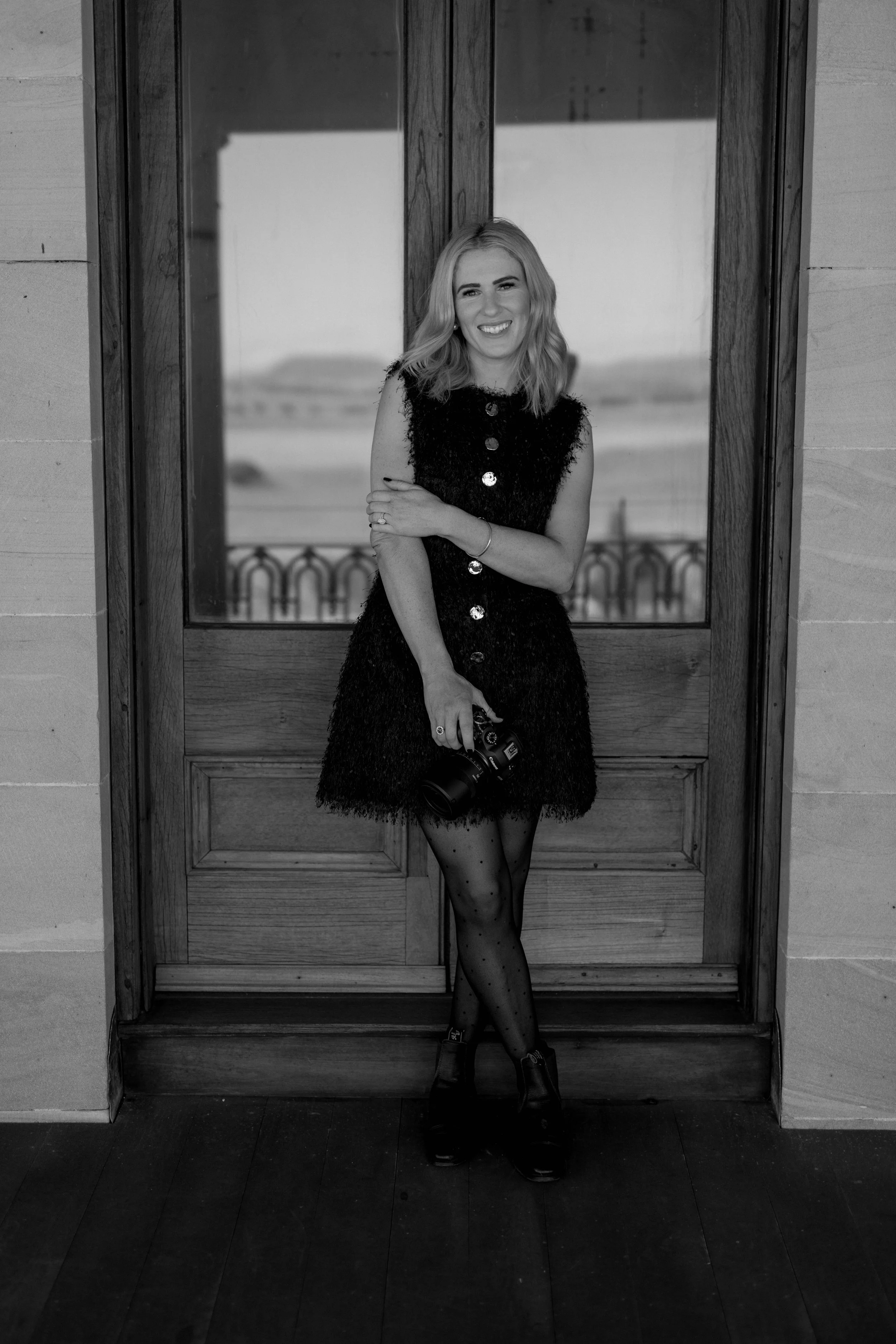 A woman standing in front of a wooden door, smiling and holding a camera, wearing a black dress and patterned tights, in a well-lit indoor space.
