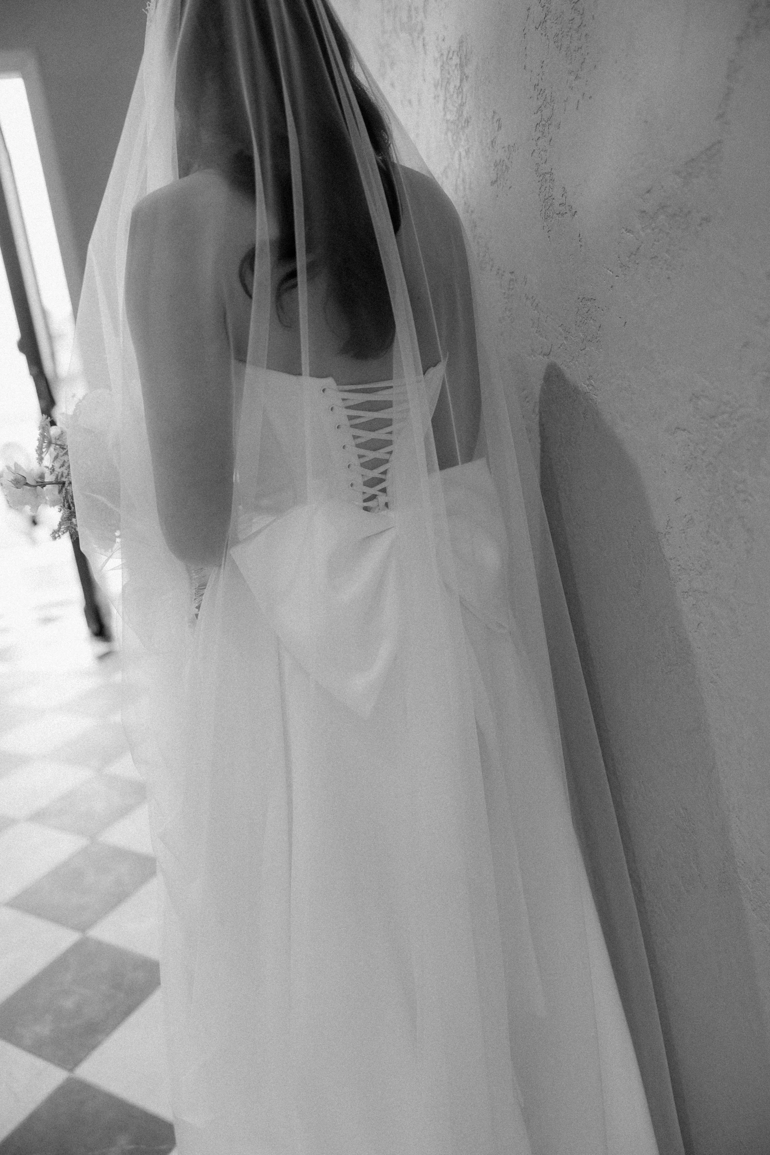 A woman in a wedding dress with a lace-up back, covered by a sheer veil, leaning against a textured wall in an indoor setting.