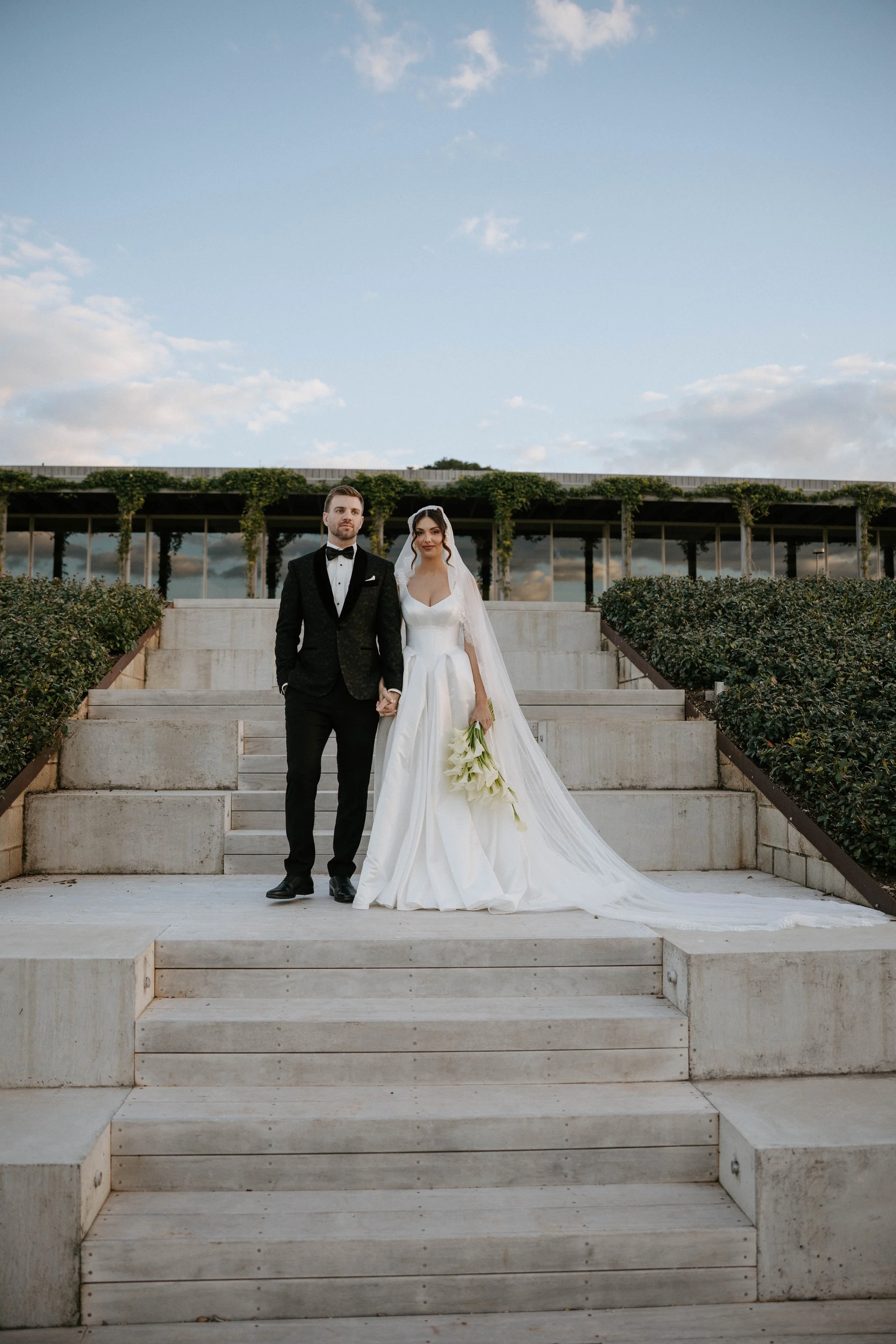 A bride and groom holding hands, standing on a staircase outdoors during daytime, with a modern building and cloudy sky behind them.