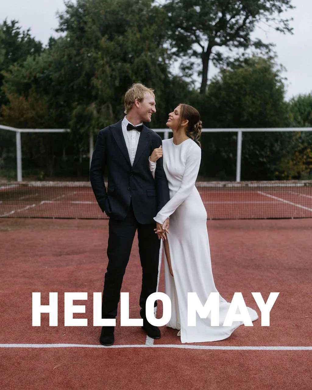 A couple in wedding attire standing on a tennis court, smiling and looking at each other. Text overlay says, "HELLO MAY."