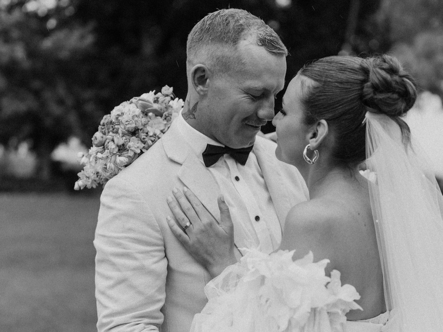 Black and white photo of a wedding couple close together, smiling with foreheads touching outdoors. The groom has short hair, a tuxedo with a bow tie, and a visible tattoo on his neck. The bride has her hair in an updo, hoop earrings, and an off-the-shoulder wedding dress. She is holding a bouquet of flowers.