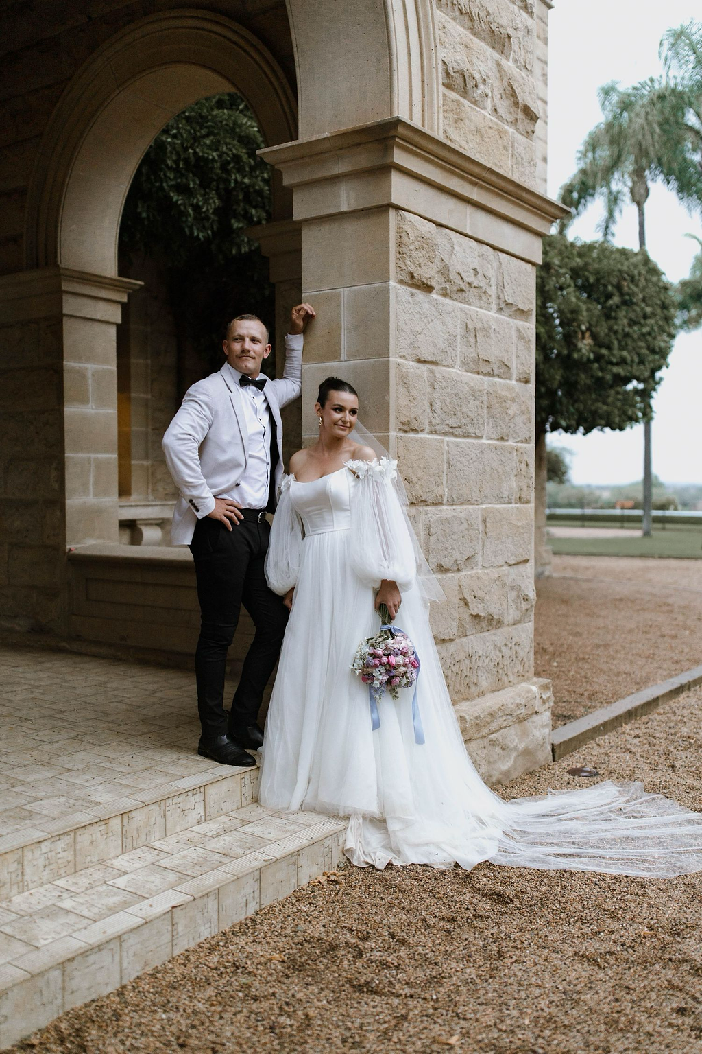 A bride in a white wedding gown holding a bouquet of pink and purple flowers, standing next to a groom in a white tuxedo jacket and black pants, outside a stone building with trees in the background.