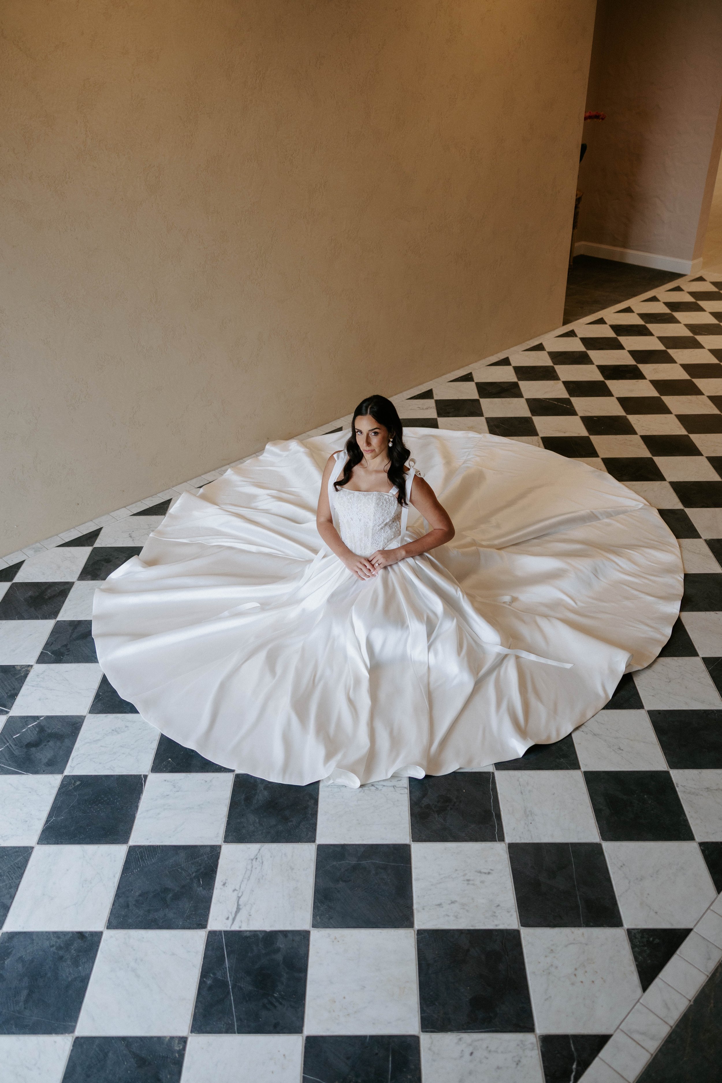 A woman in a white wedding dress sitting on the floor with her hands in her lap, surrounded by her dress's long train, in a room with beige walls and black-and-white checkered tile flooring.