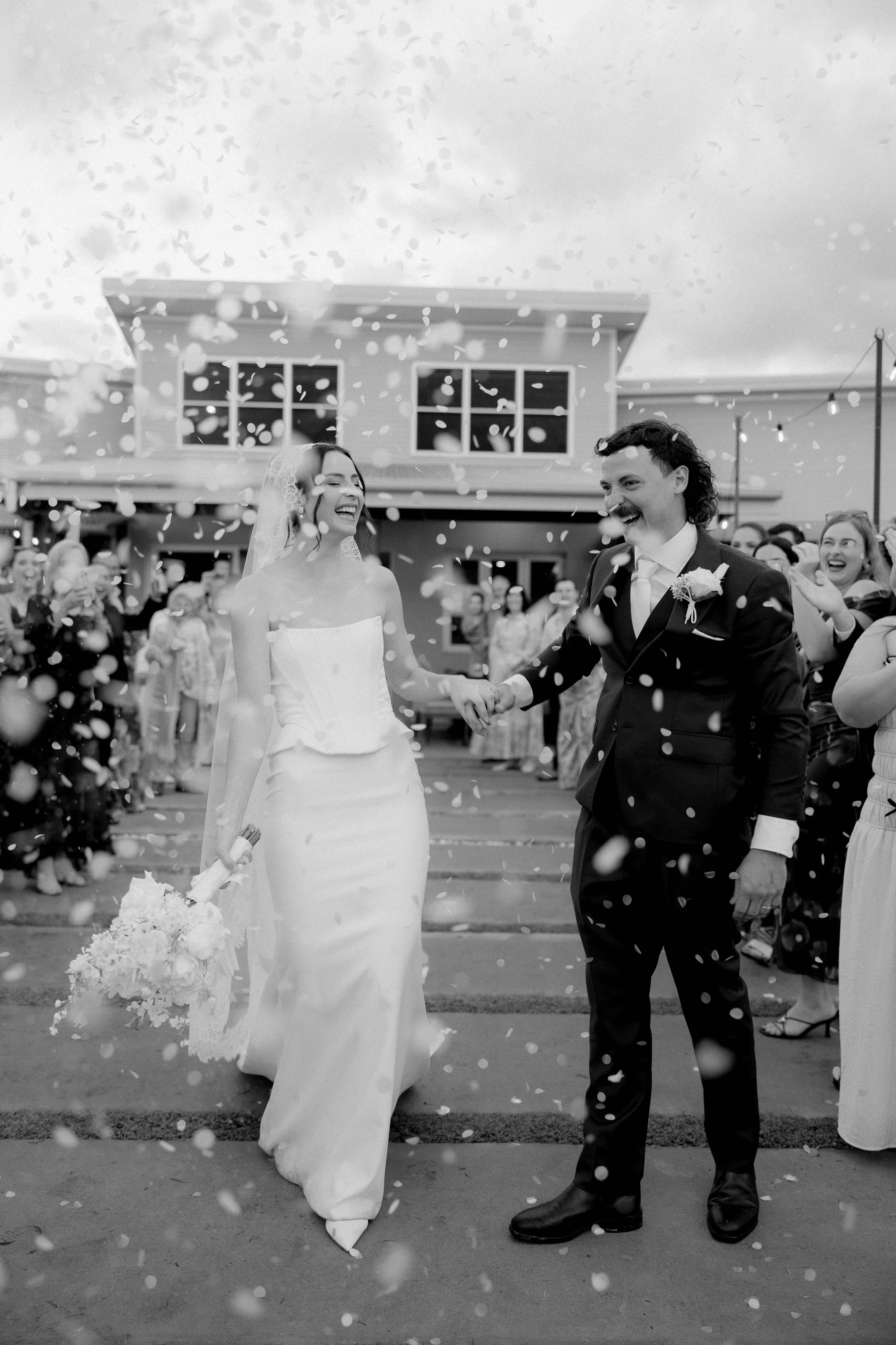 A black-and-white photo of a bride and groom smiling as they walk hand-in-hand. The bride wears a white strapless wedding dress and veil, holding a bouquet of flowers. The groom wears a dark suit with a boutonniere. They are surrounded by guests, with confetti falling around them, celebrating their wedding outdoors in front of a building.