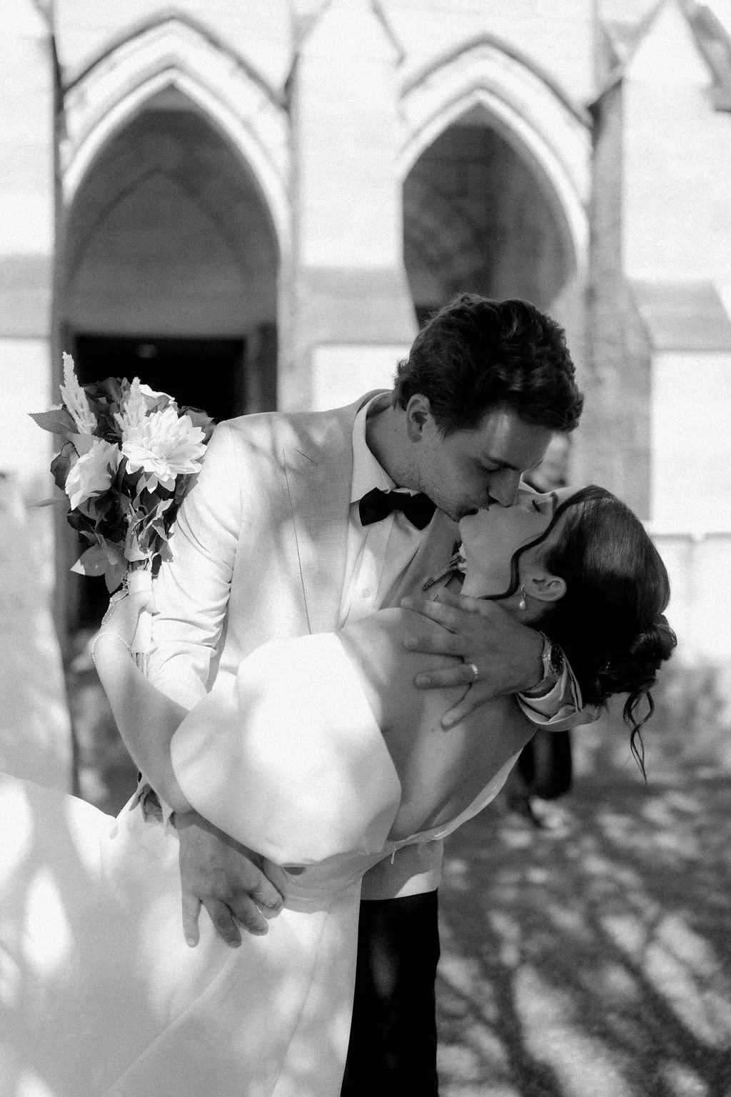 A black and white photo of a couple sharing a kiss outside a building with arched windows, the woman is in a white dress and the man in a white tuxedo with a bow tie.