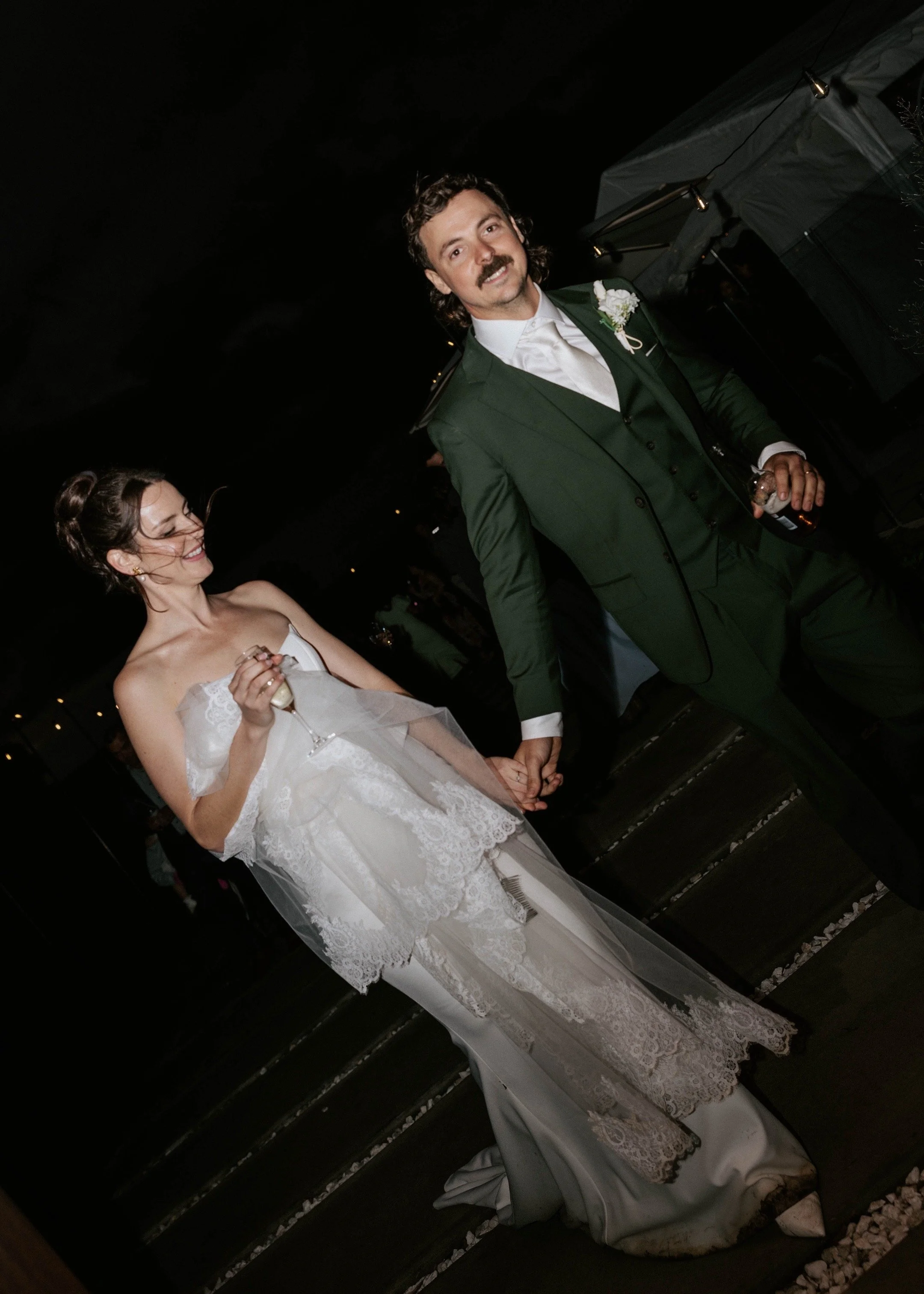 A bride and groom holding hands at night, the bride in a white wedding dress and veil, the groom in a green suit, both smiling.