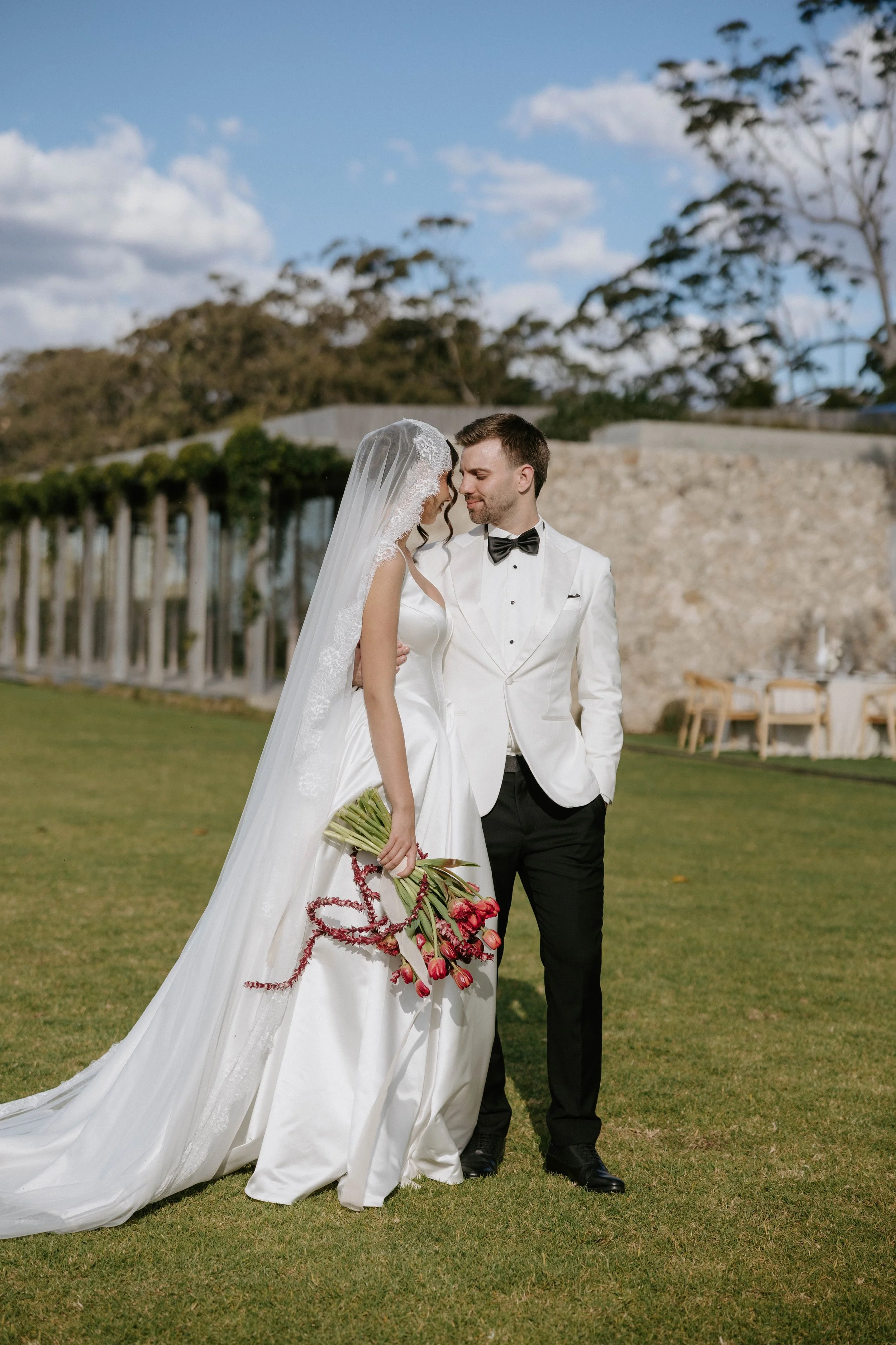A bride and groom standing close together outdoors, with the bride holding a bouquet of pink and red tulips. The bride wears a long white wedding gown and veil, and the groom wears a white tuxedo jacket with black pants and a black bow tie. They are 