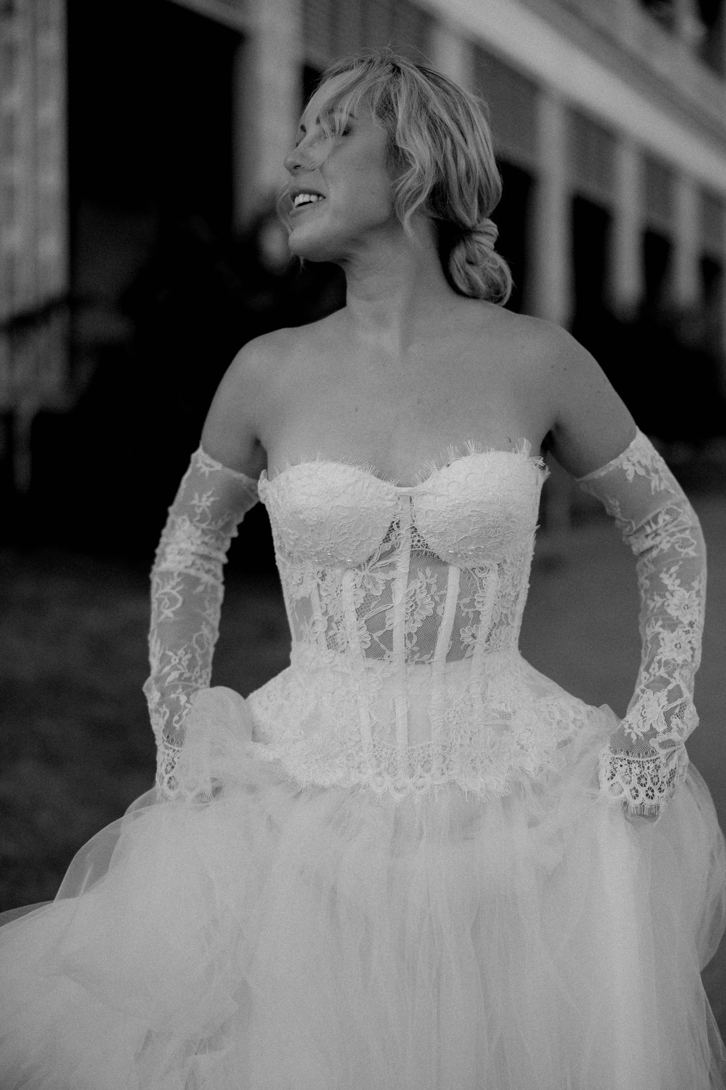 Black and white photo of a woman wearing a lace and tulle wedding dress, smiling with her eyes closed, outdoors in front of a building.
