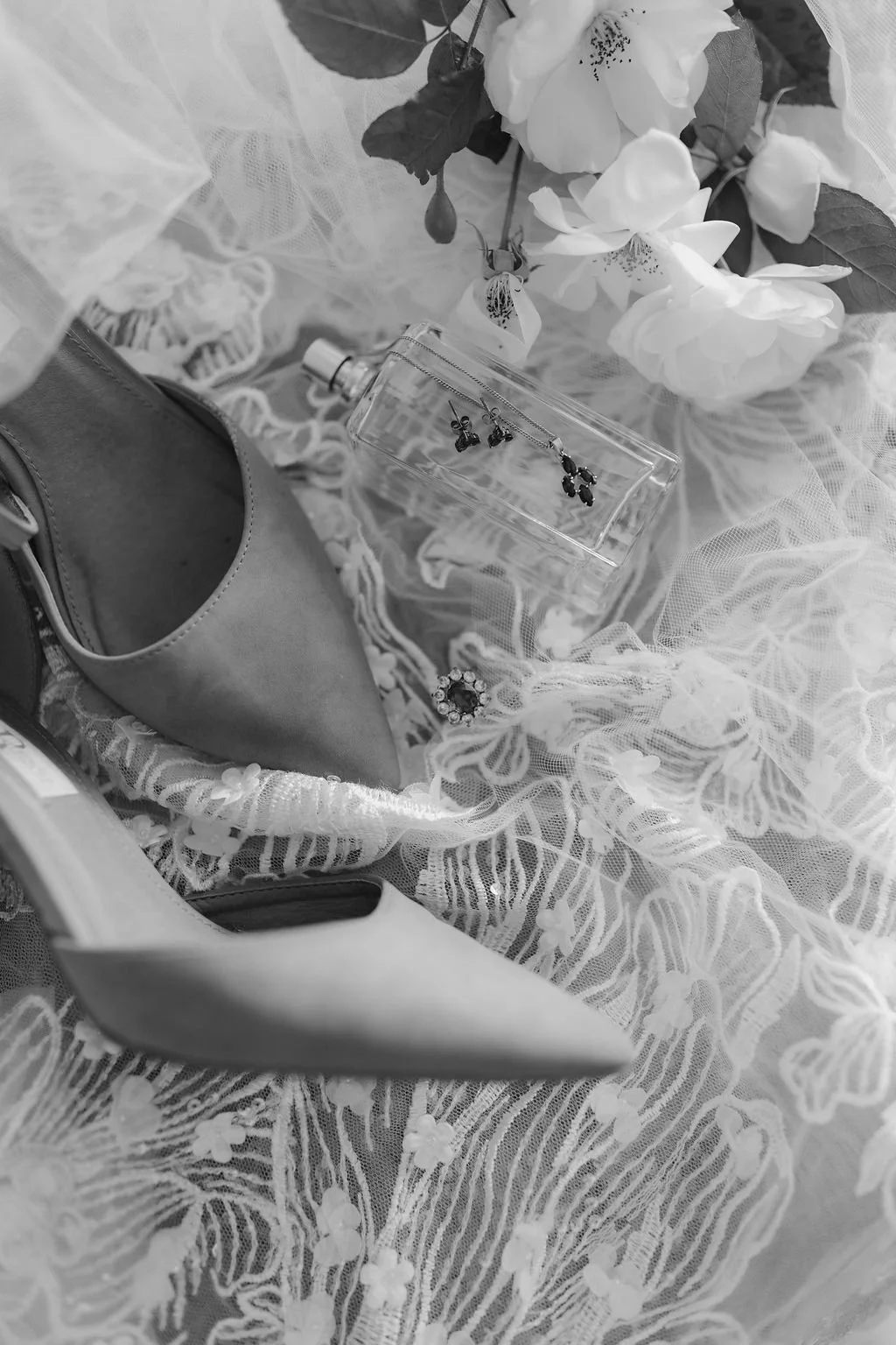 A collection of jewelry including earrings, a ring, and necklaces displayed on lace fabric, with flowers, a perfume bottle, a bag, and a shoe surrounding them.