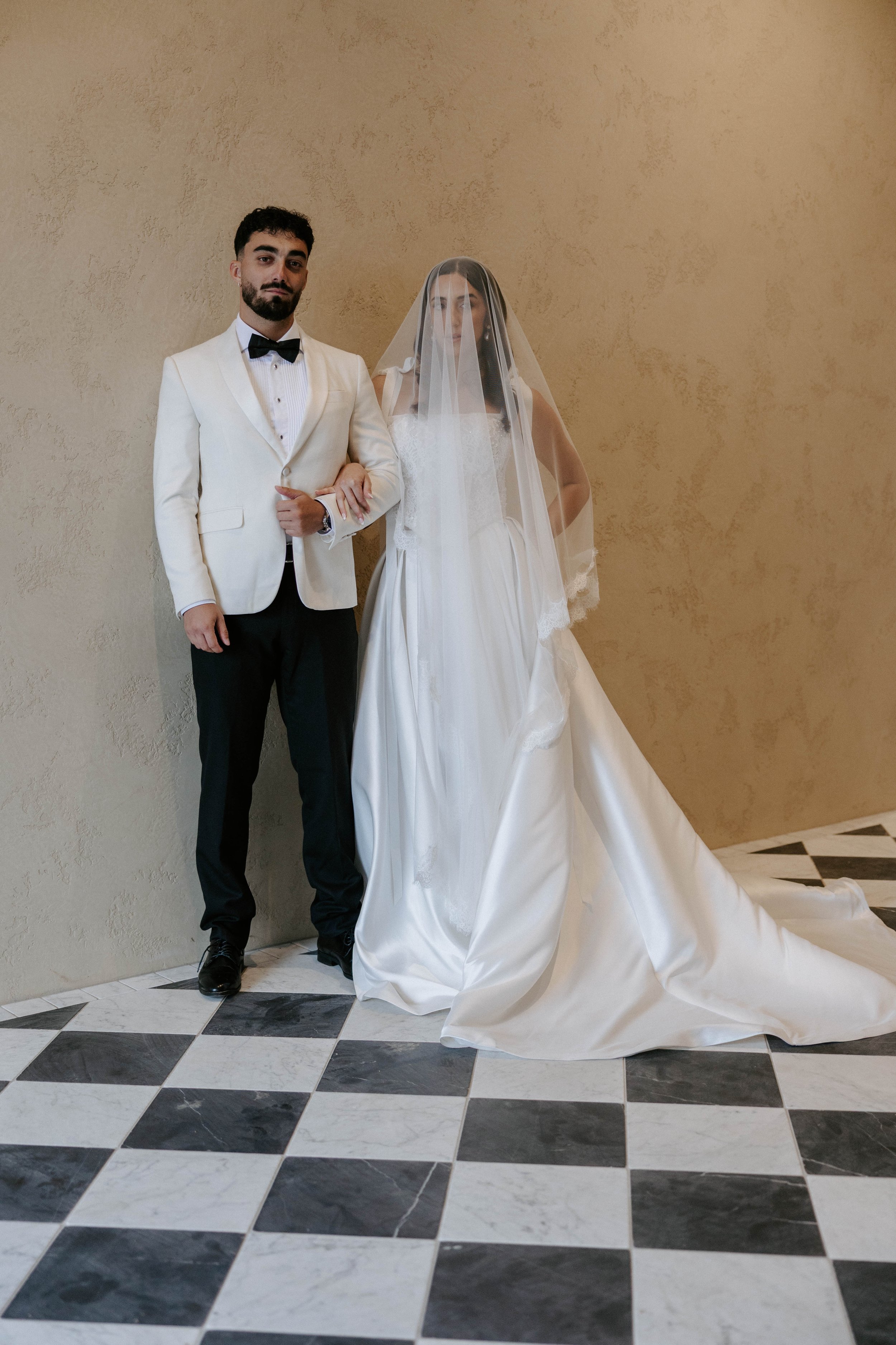 A groom and bride standing together in wedding attire against a beige wall, with black and white checkered floor tiles.