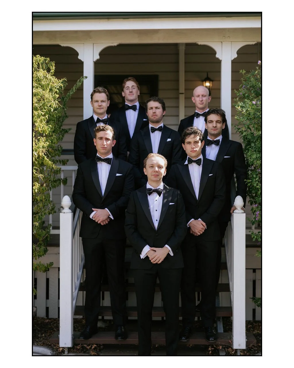 Group of nine men in tuxedos standing on front porch of house, posing for a formal photo.
