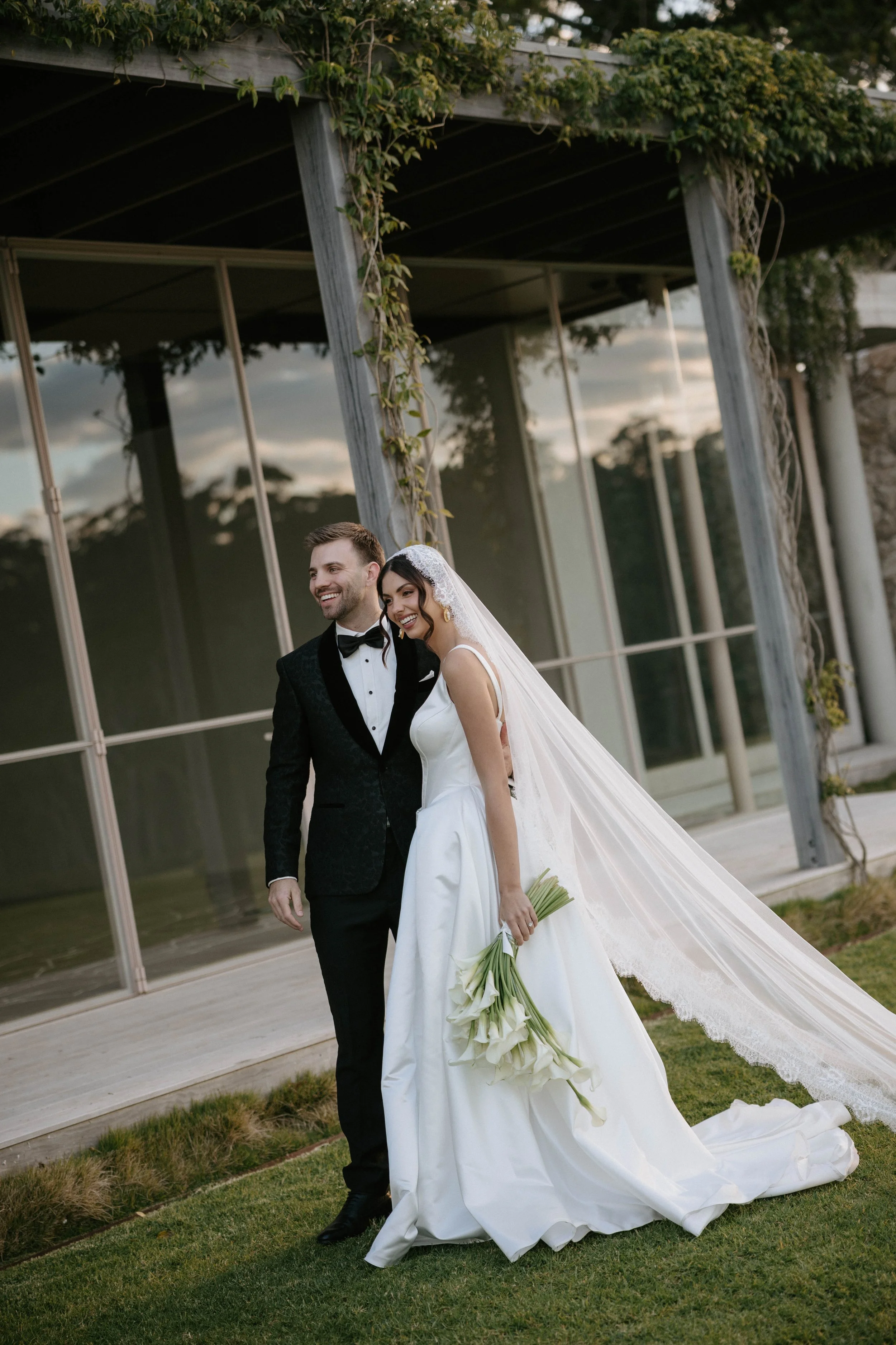 A happy bride and groom walking outdoors near a modern building with glass windows, the bride holding a bouquet of white calla lilies and wearing a white wedding gown with a long train, the groom dressed in a black tuxedo with a bowtie.
