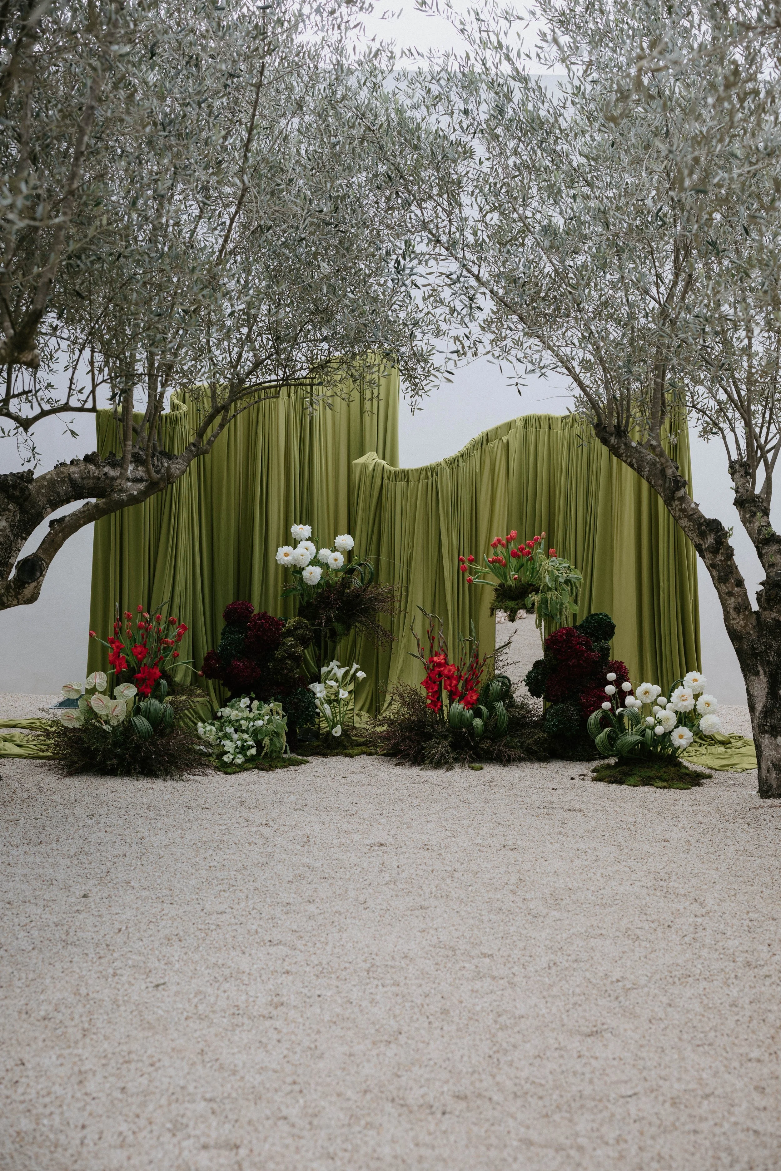 Indoor wedding altar with green drapery backdrop, surrounded by potted plants and flowers, with trees framing the scene.
