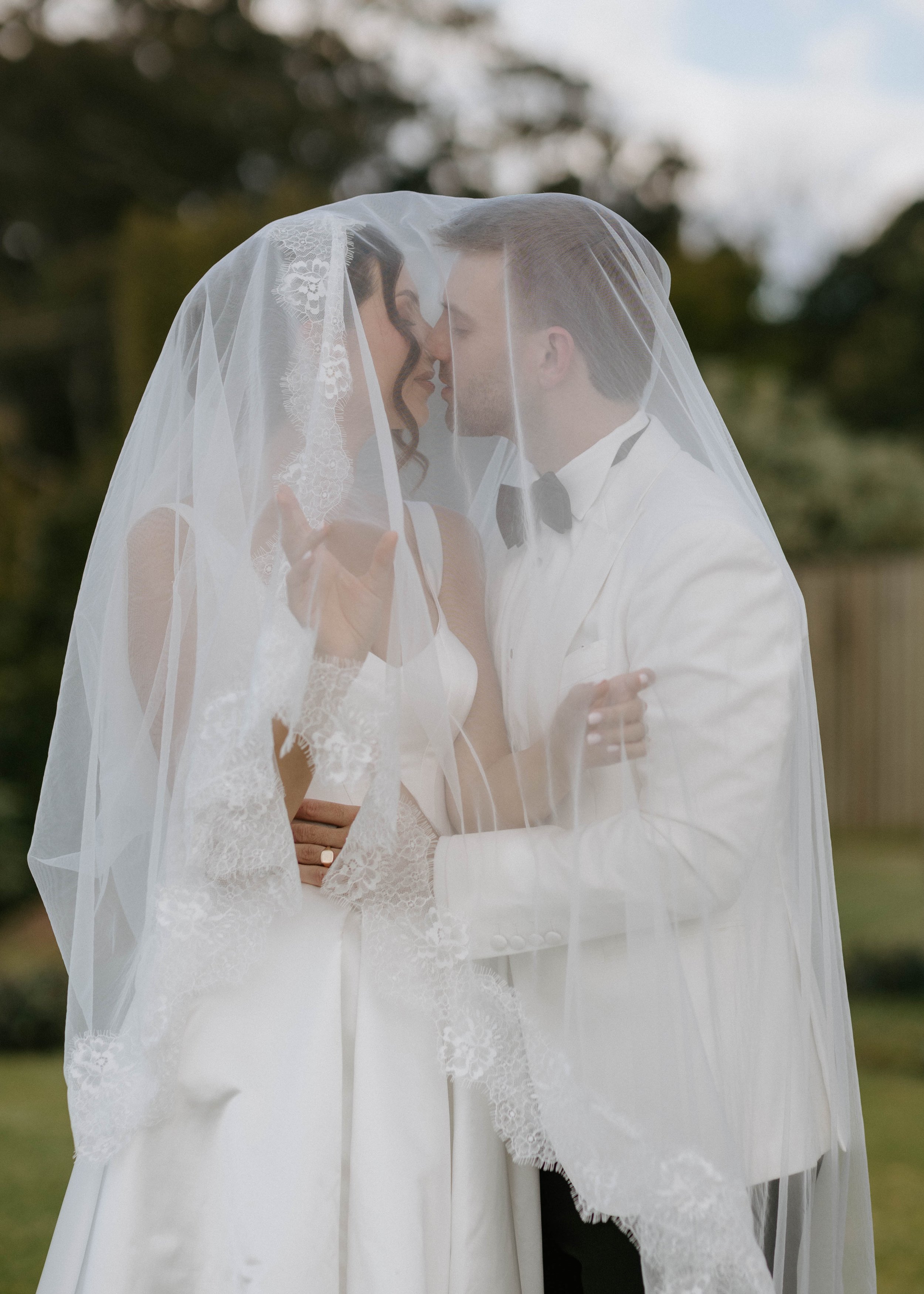 A bride and groom dressed in wedding attire share a kiss beneath a lace veil outdoors.