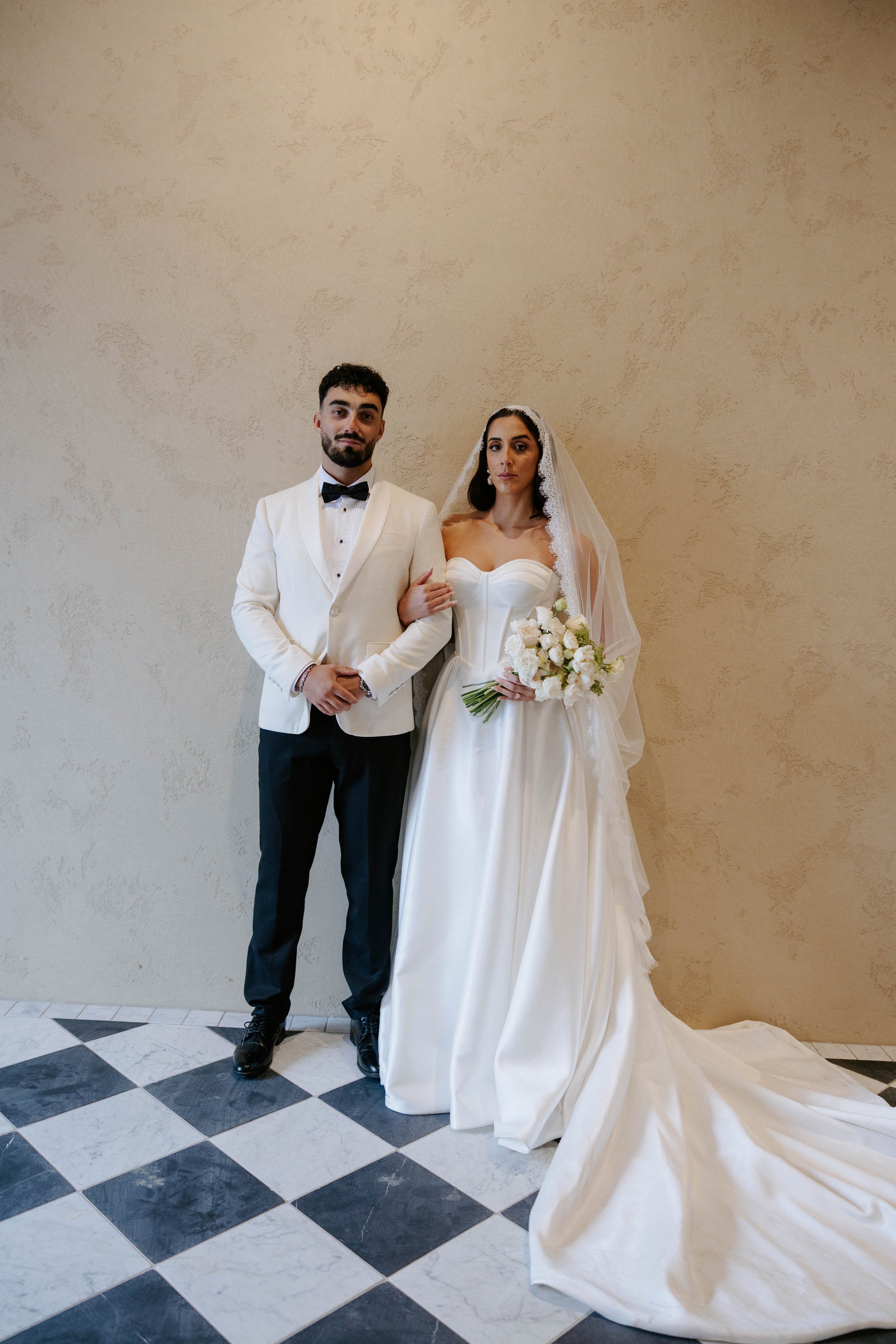 A groom in a tuxedo and a bride in a wedding dress holding a bouquet pose against a beige wall with a checkered black and white tile floor.