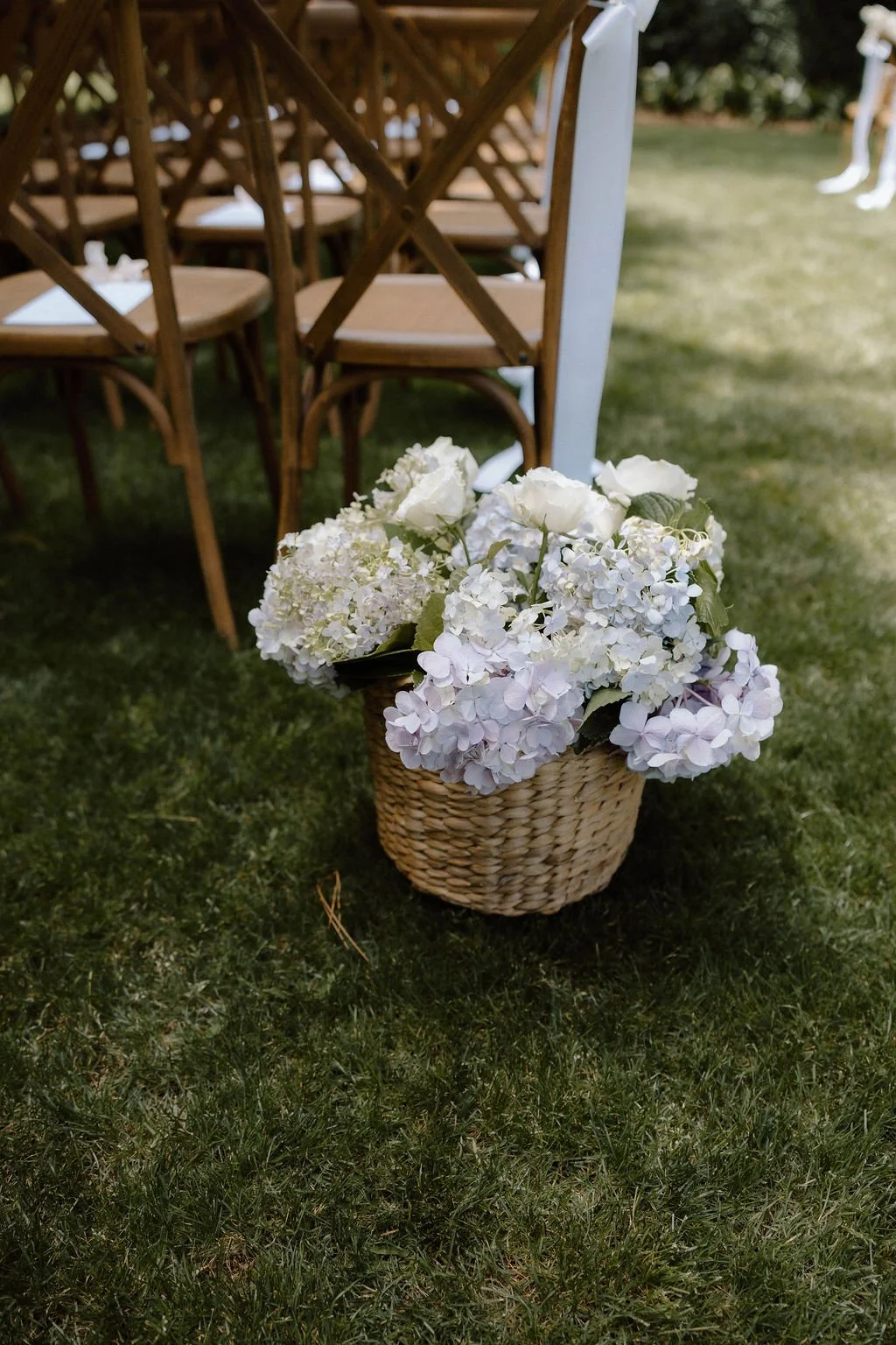 A woven basket filled with white and light purple hydrangeas and white roses, placed on grass next to wooden chairs and a white table at an outdoor event.