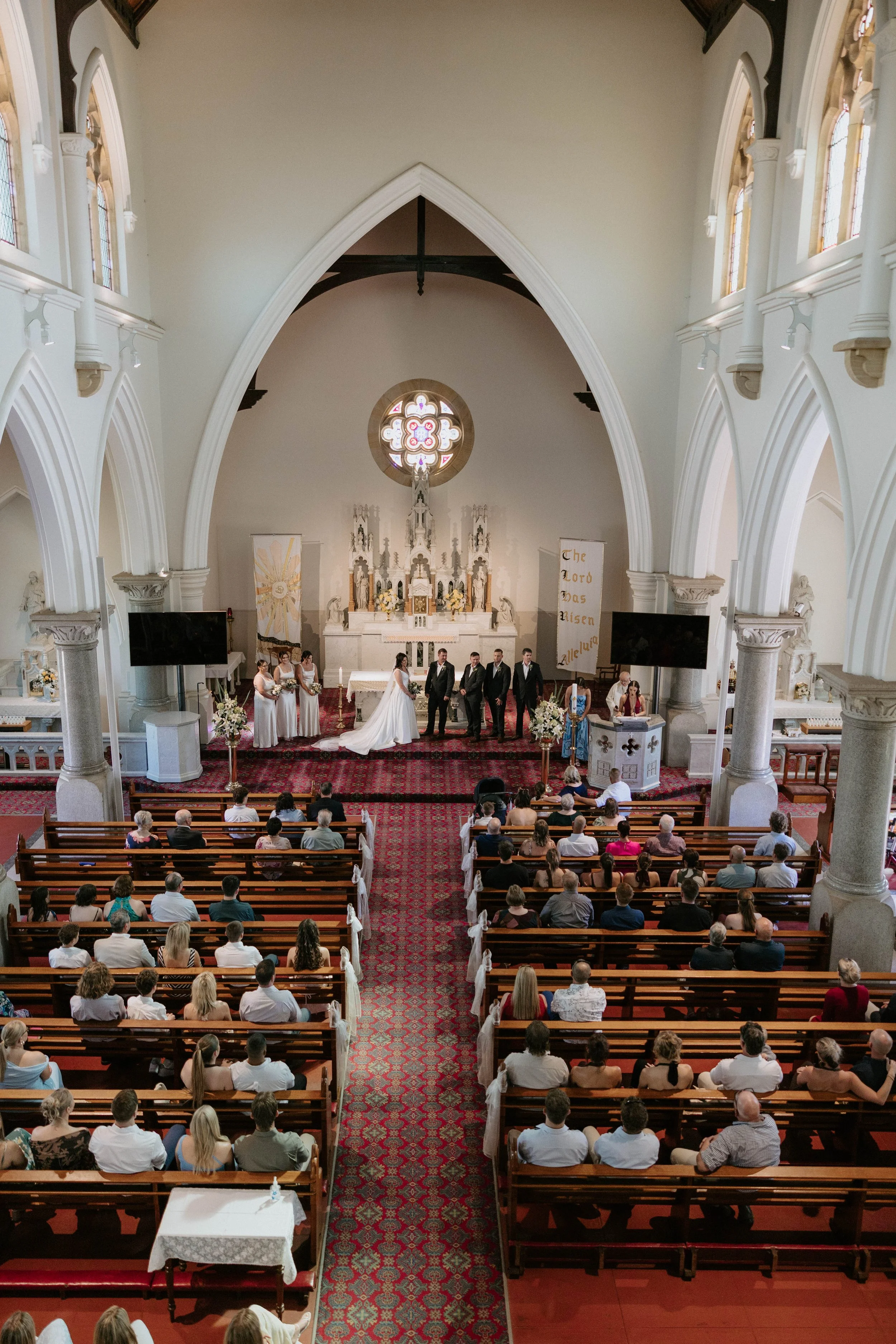 Inside a church during a wedding ceremony, with rows of pews filled with guests, an altar at the front featuring a bride and groom, bridesmaids, groomsmen, and worship decorations, stained glass windows, and religious banners.