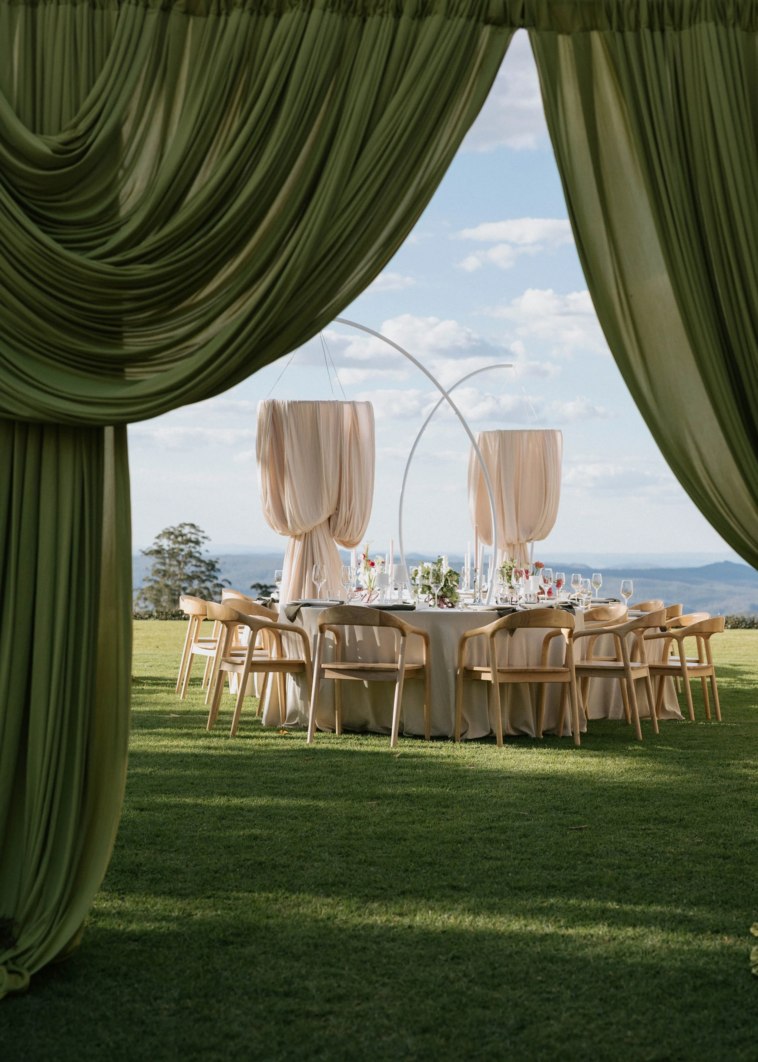 A decorated outdoor banquet table set up for a wedding reception or special event, viewed through green curtains. The table is draped with a white tablecloth, surrounded by wooden chairs, and features floral arrangements and glassware. Large beige fa