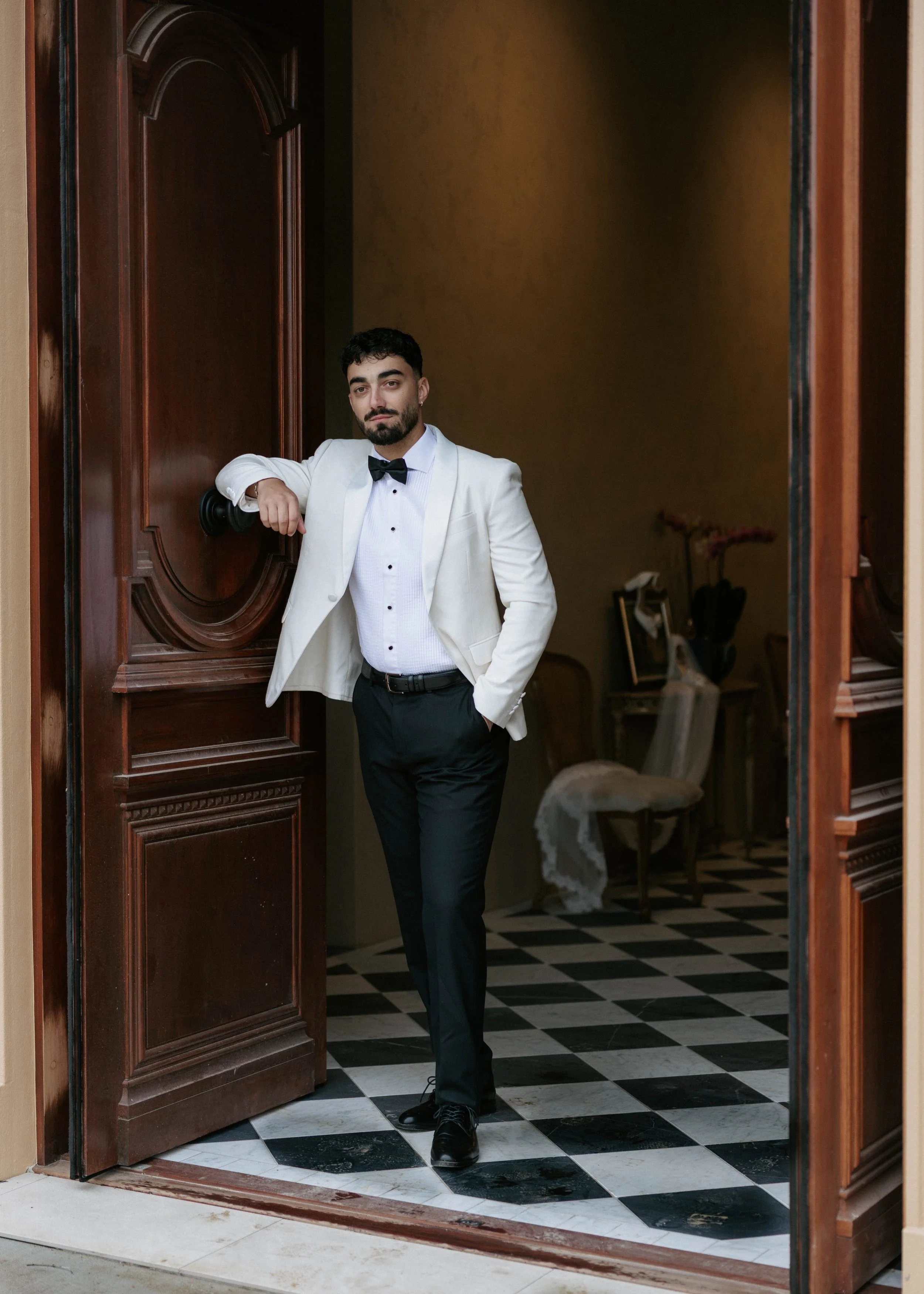 A man in a white tuxedo jacket, black bow tie, and black pants leaning casually against a wooden doorframe in a room with checkered black and white tile flooring and a beige wall.