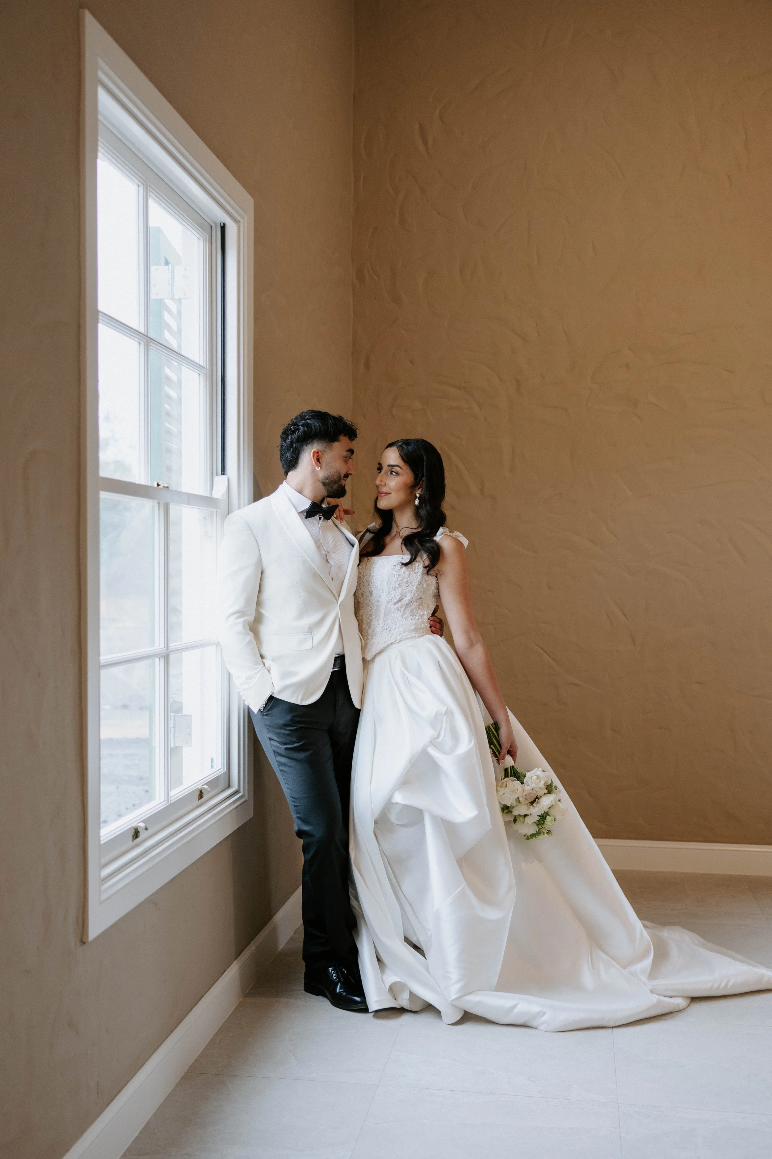 A bride and groom standing close together by a window, sharing a tender moment, with the bride holding a bouquet of white roses.