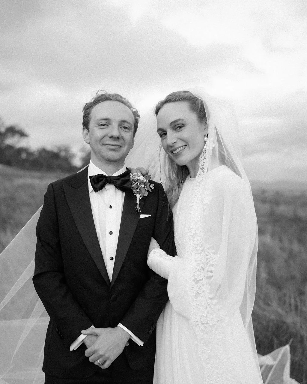 Black and white photo of a bride and groom standing outdoors, with the bride holding the groom's arm. The groom is wearing a tuxedo with a bow tie and boutonniere, and the bride is wearing a long-sleeved lace wedding dress with a veil. They are smili