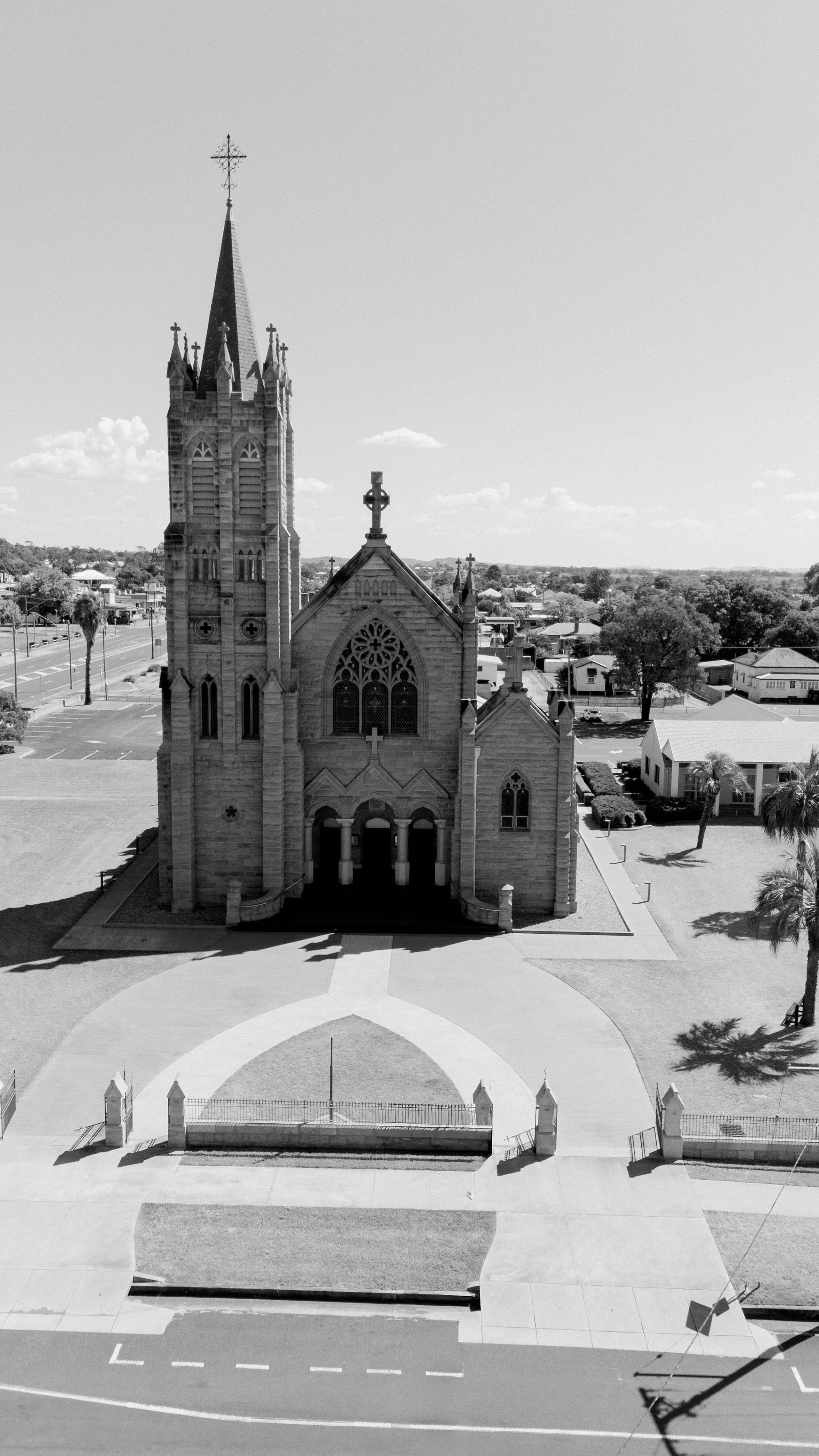 Black and white photo of a historic stone church with a tall steeple, large stained glass windows, and religious crosses on its roof, surrounded by a well-maintained lawn, trees, and nearby buildings in a suburban area.