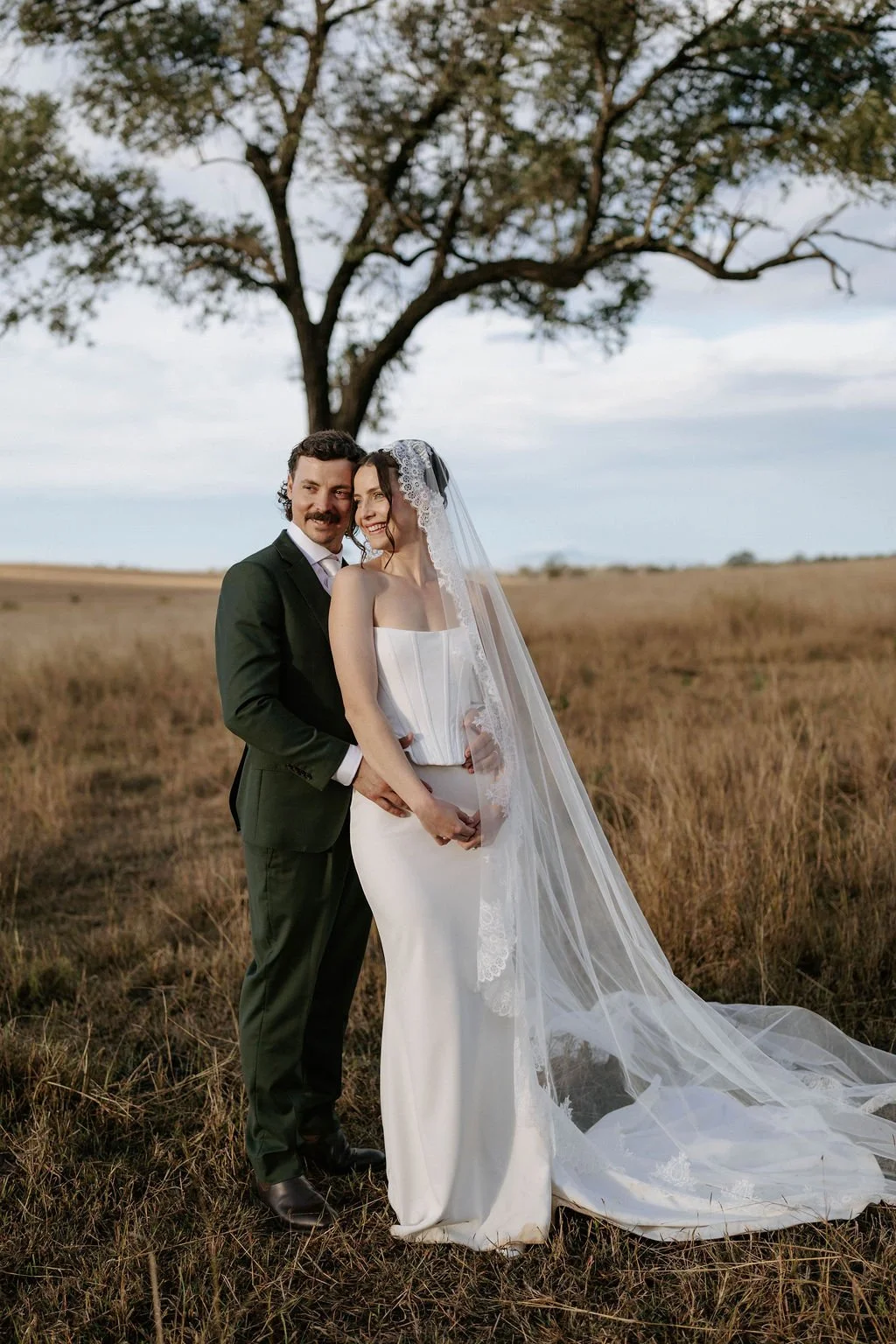 A newlywed couple standing in a field with a large tree in the background, smiling and embracing each other. The bride wears a white strapless wedding gown and veil, while the groom is in a dark suit with a white shirt and tie.