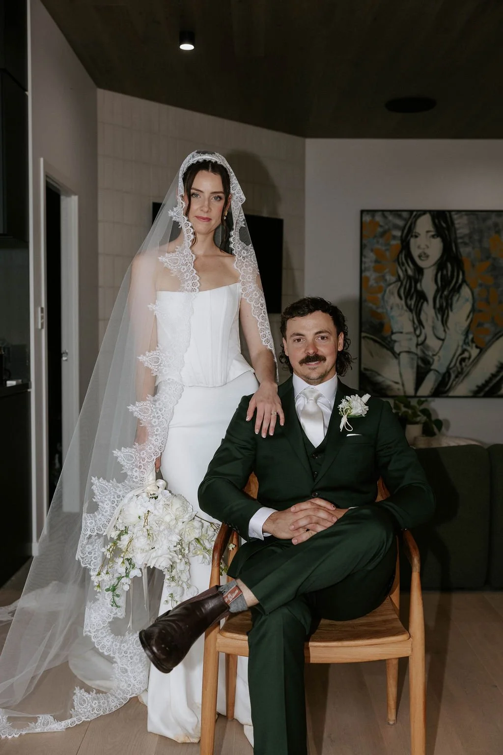 A bride in a white wedding dress with a lace veil stands behind a seated groom in a dark green suit, inside a modern room with art on the wall.