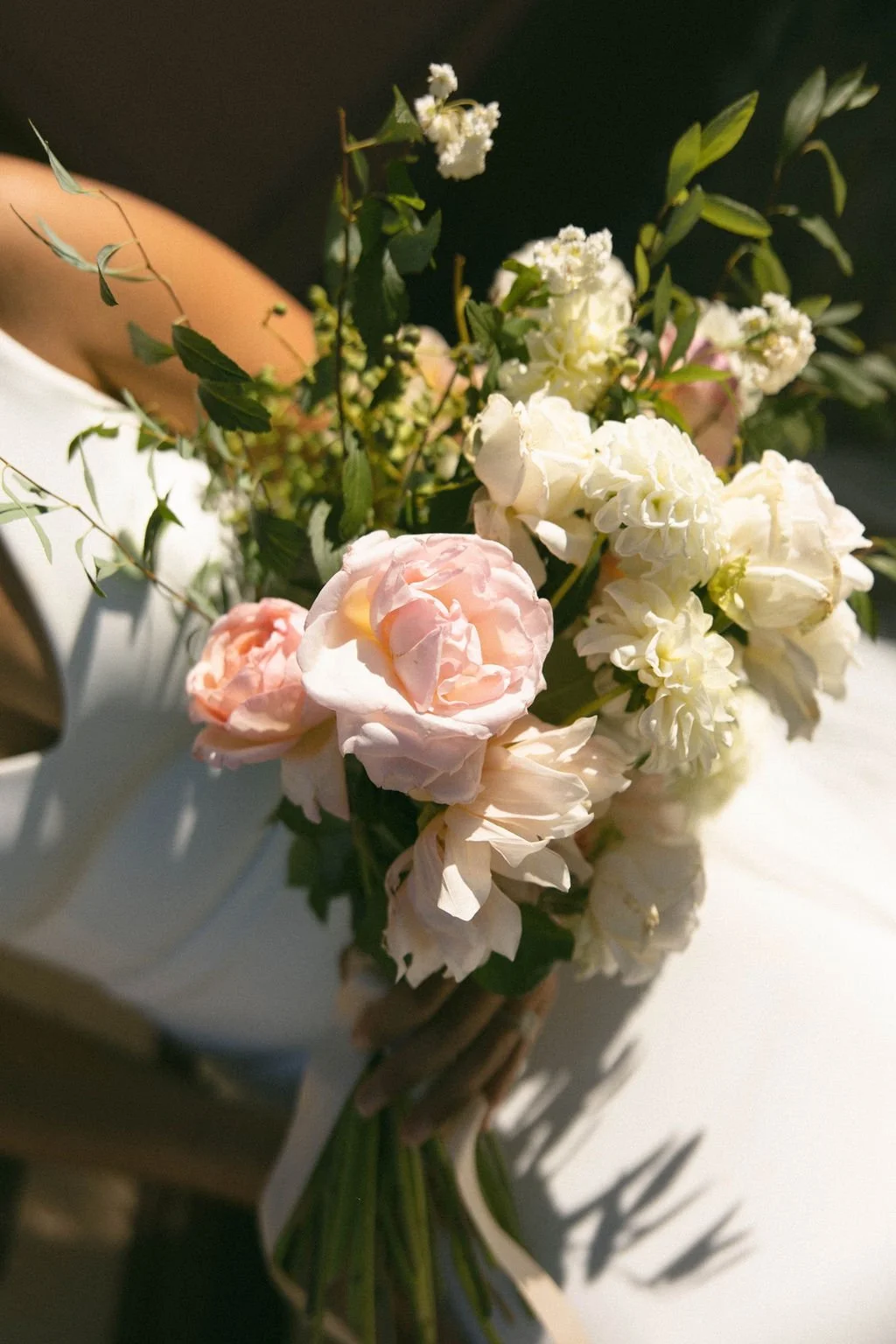 A bouquet of white and pale pink flowers with green leaves on a white surface, with sunlight casting shadows.