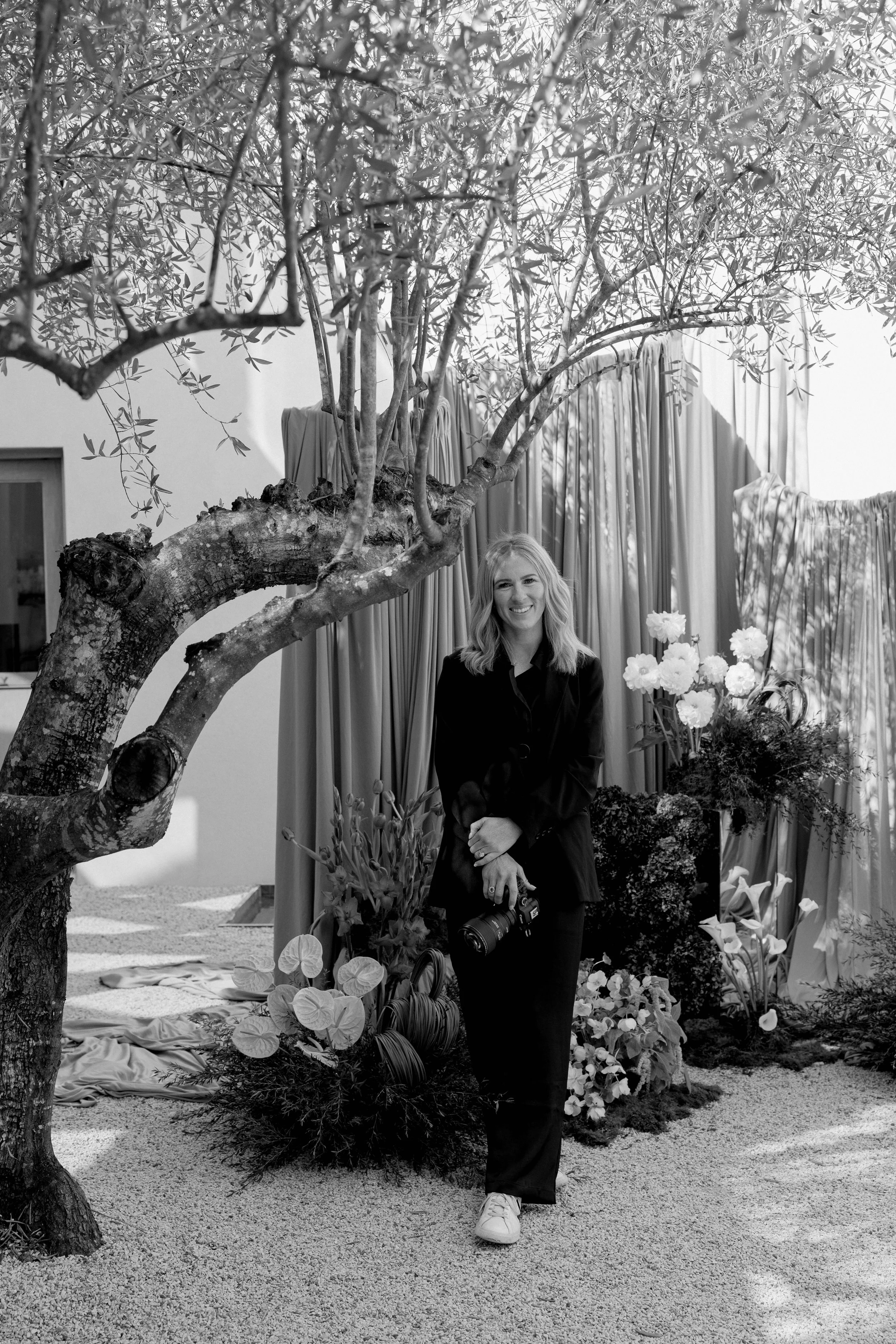 A woman standing outdoors among plants and flowers, holding a camera, smiling, with a large tree and curtains in the background, in black and white.