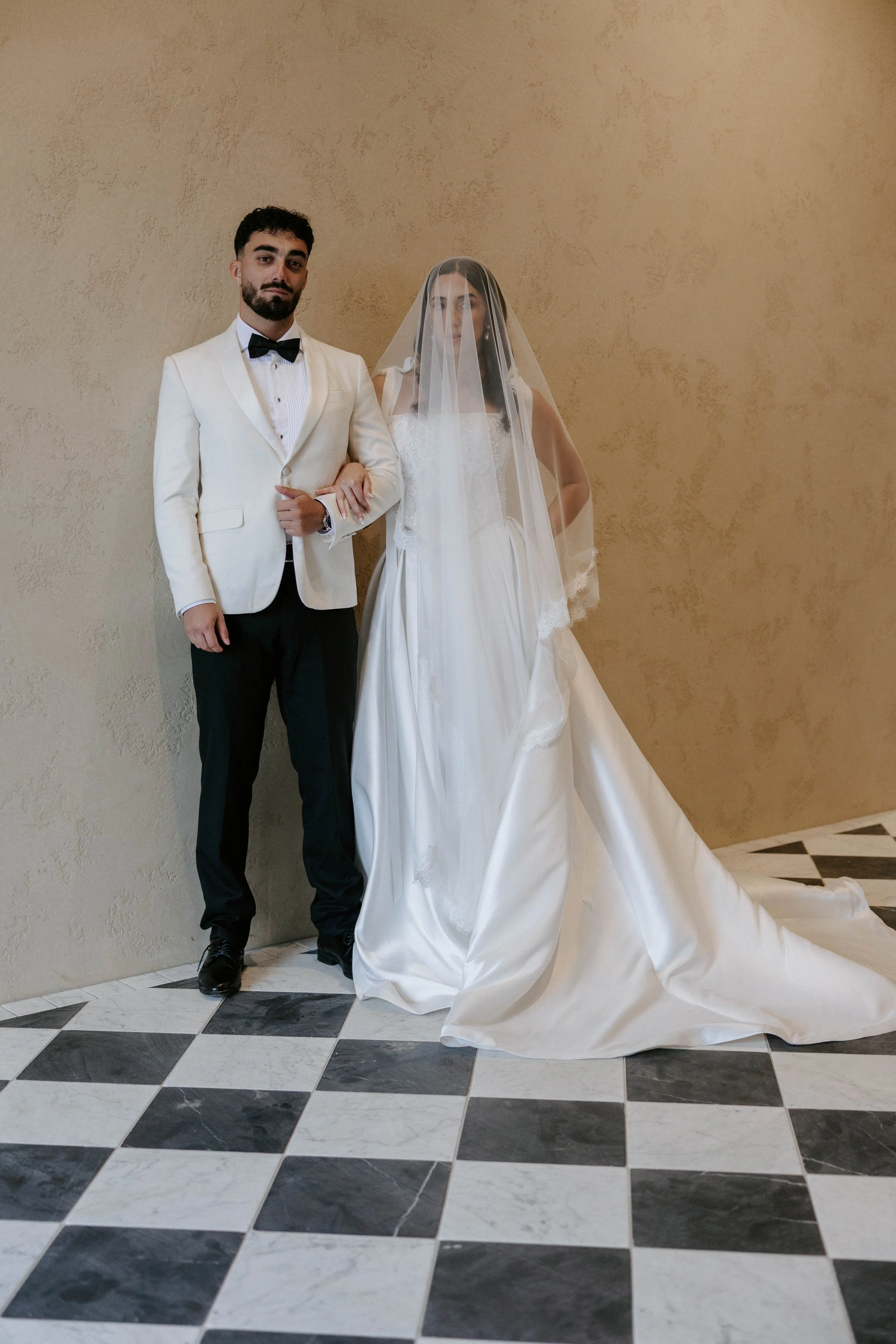 A man in a cream tuxedo jacket with a bow tie standing next to a woman in a wedding dress with a veil, both posing inside a building with beige walls and black-and-white checkered tile flooring.