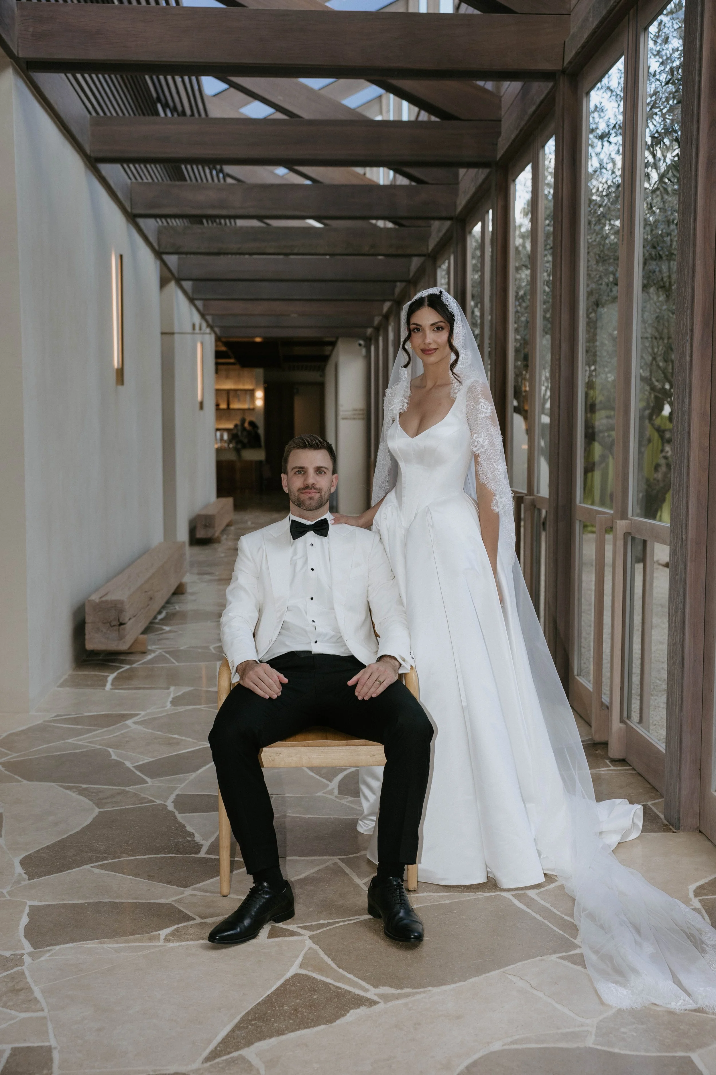 A bride in a white wedding gown with a lace veil stands behind a seated groom in a tuxedo, inside a modern building with large windows and stone flooring.