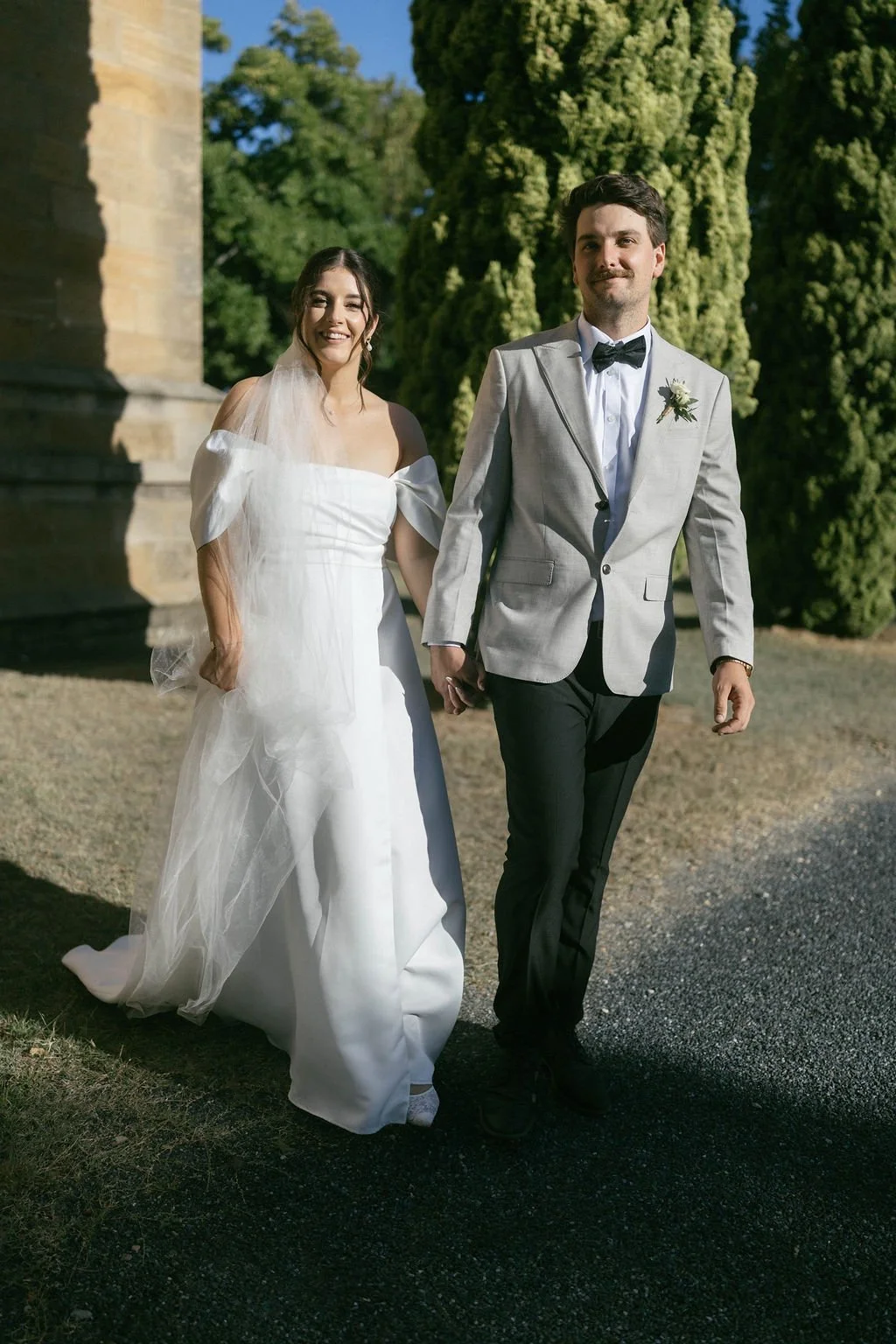 A bride and groom walking outdoors on a sunny day, holding hands, smiling, with green trees in the background.